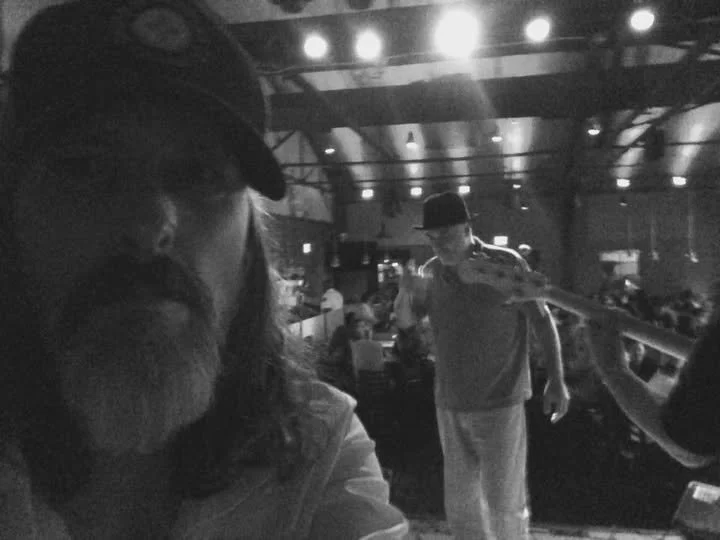 A black and white photo of a man with long hair and a beard wearing a cap in a restaurant or bar. In the background, a person wearing a hat is receiving a drink from a server, with other patrons seated at tables.