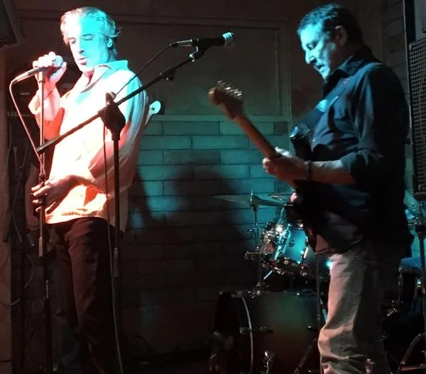 The ZigZags performing on stage at Tiny's in Santa Fe, musicians, Henry Sutro and Mike Montiel,  with a drum set in the background, one singing into a microphone and the other playing an electric guitar, with a brick wall backdrop.
