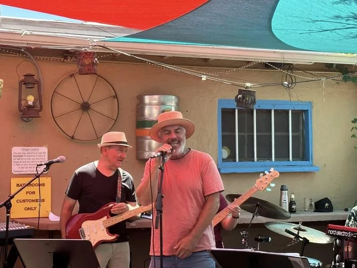 Henry Sutro performing music outdoors at Cowgirl Hall of Fame in Santa Fe, NM, one singing with a microphone and the other playing an electric guitar, under colorful shade cloths with a wall decorated with vintage items.