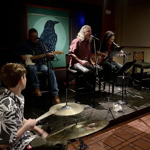 The Nuevo Hippies led by singer Henry Sutro at El Farol in Santa Fe, NM, performing on stage with a young drummer in the foreground and a large bird illustration in the background.