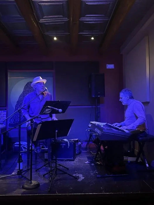 Two musicians performing on a stage in a cozy, dimly lit venue. One is singing into a microphone while wearing a hat, and the other is playing a keyboard. The background includes a large artwork and dark wood paneling.