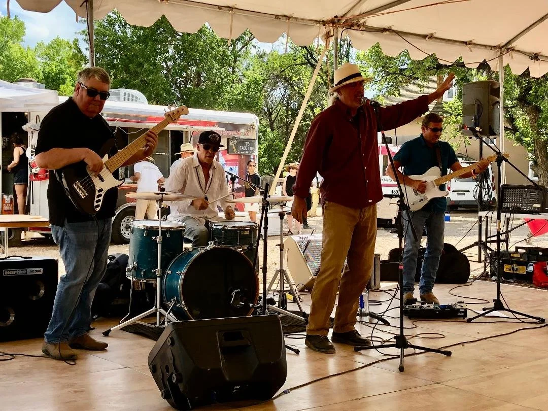 A four-piece band performing on an outdoor stage under a tent, with trees and food trucks in the background. The band includes a guitarist, a drummer, a vocalist, and another guitarist, all casually dressed. The vocalist is wearing a hat and singing 