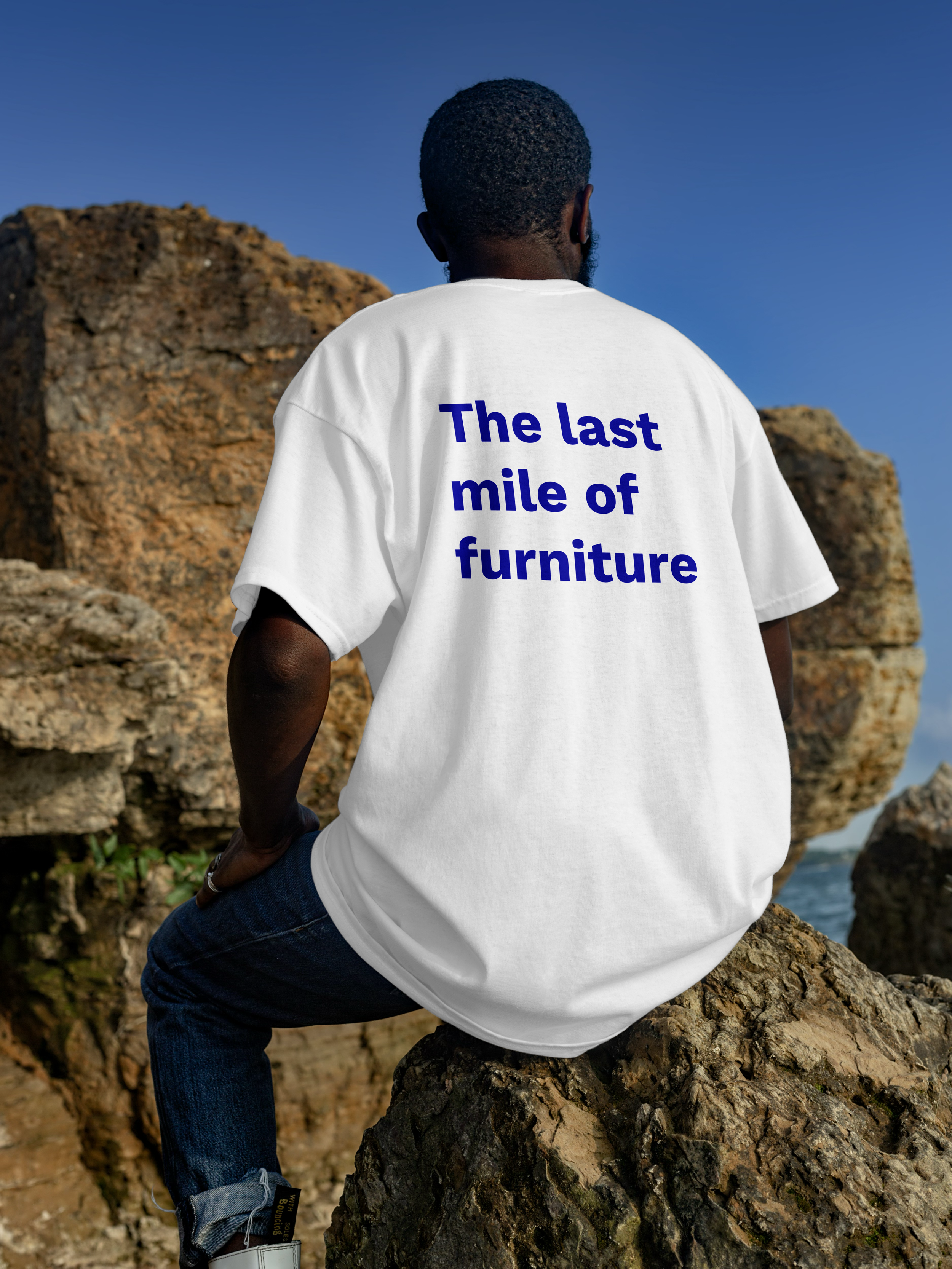 Un uomo con la schiena rivolta che indossa una maglietta bianca con la scritta blu 'The last mile of furniture' e si trova su una roccia sulla spiaggia, con un cielo azzurro sullo sfondo.