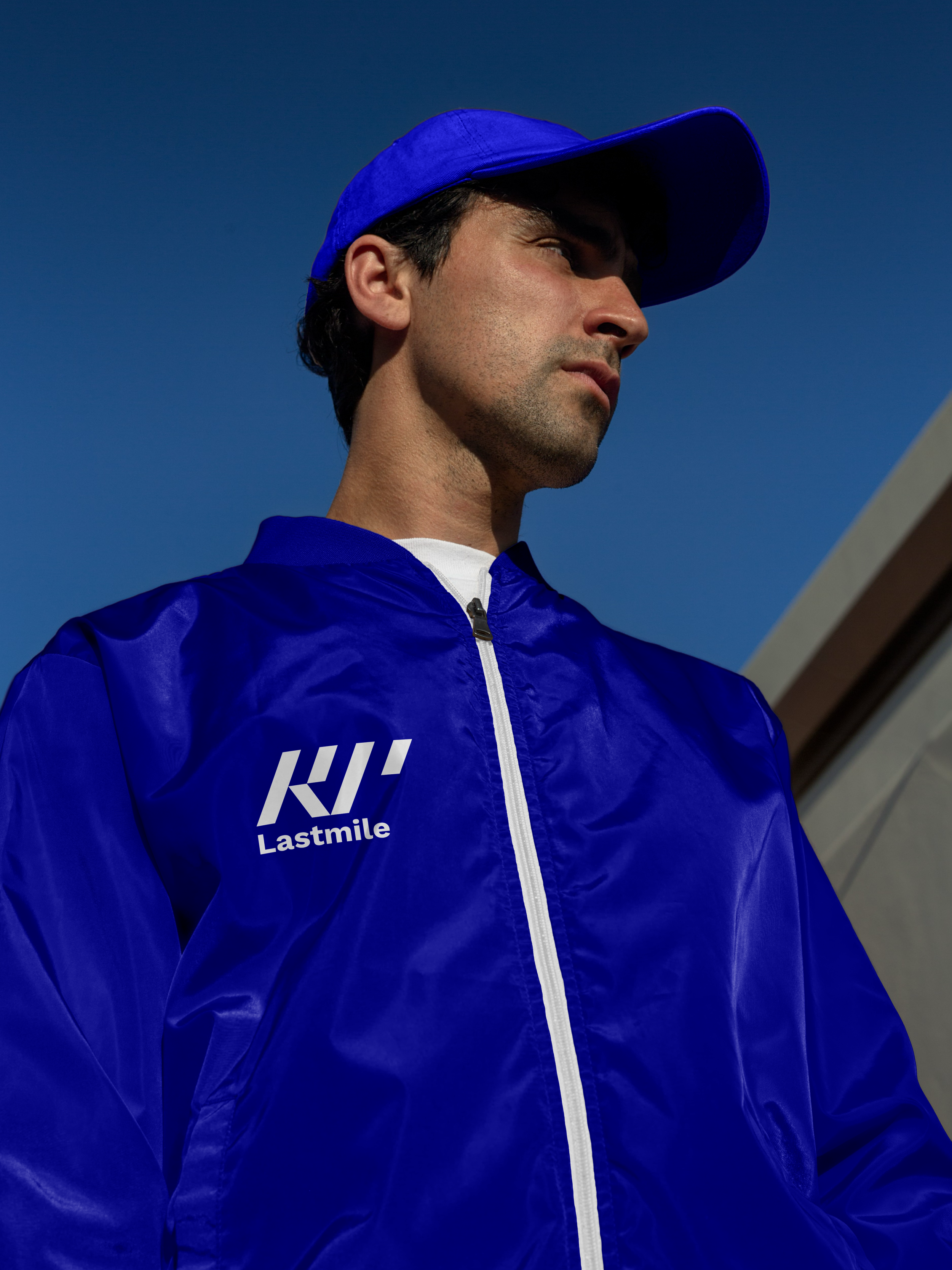 Giovane uomo con cappellino blu e giacca blu con logo 'Lastmile' scritto sopra, vista laterale.