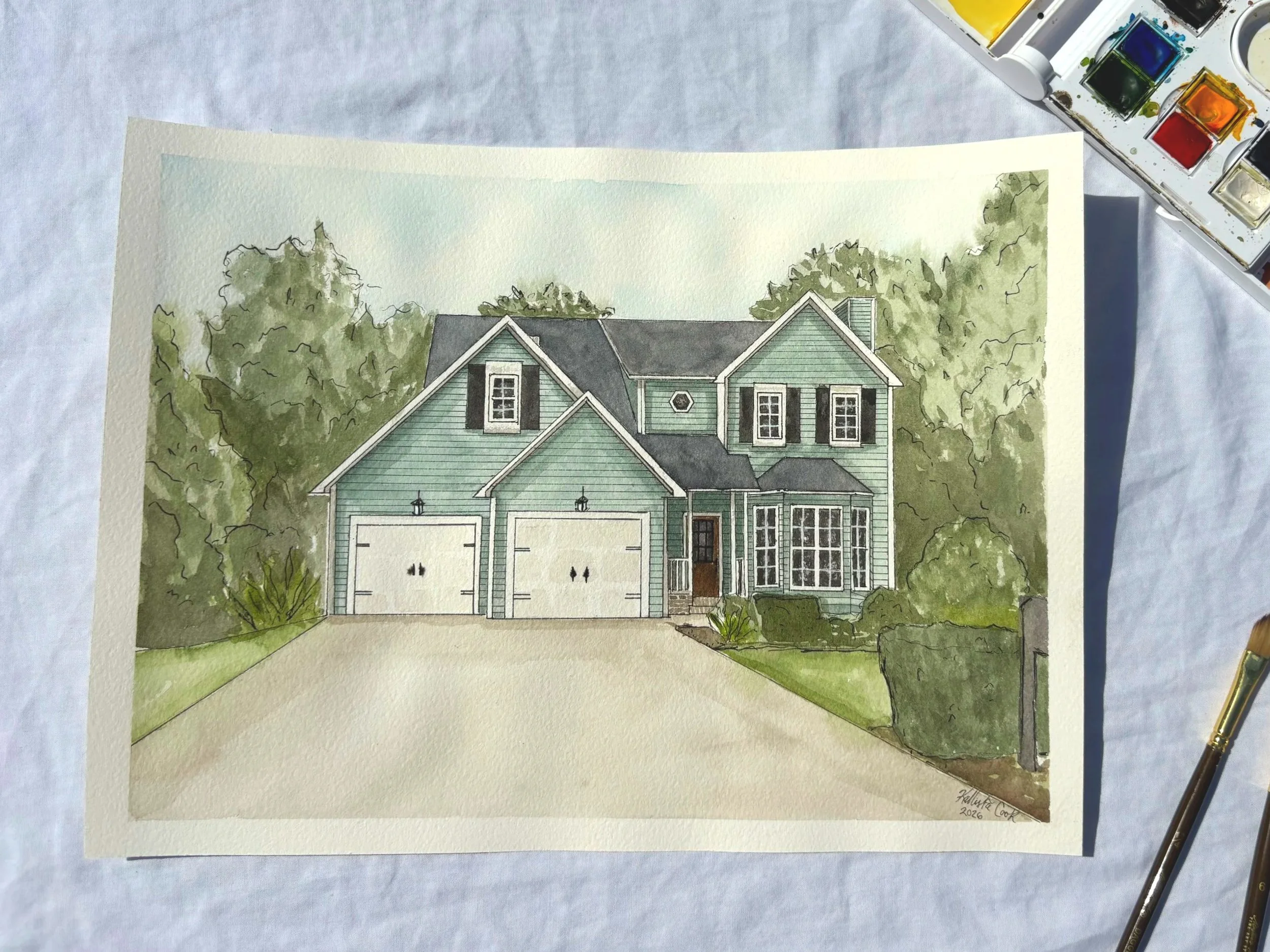 Watercolor painting of a two-story house with a green exterior, black shutters, and a garage, surrounded by trees and bushes, displayed on a white surface with watercolor paints and a paintbrush nearby.