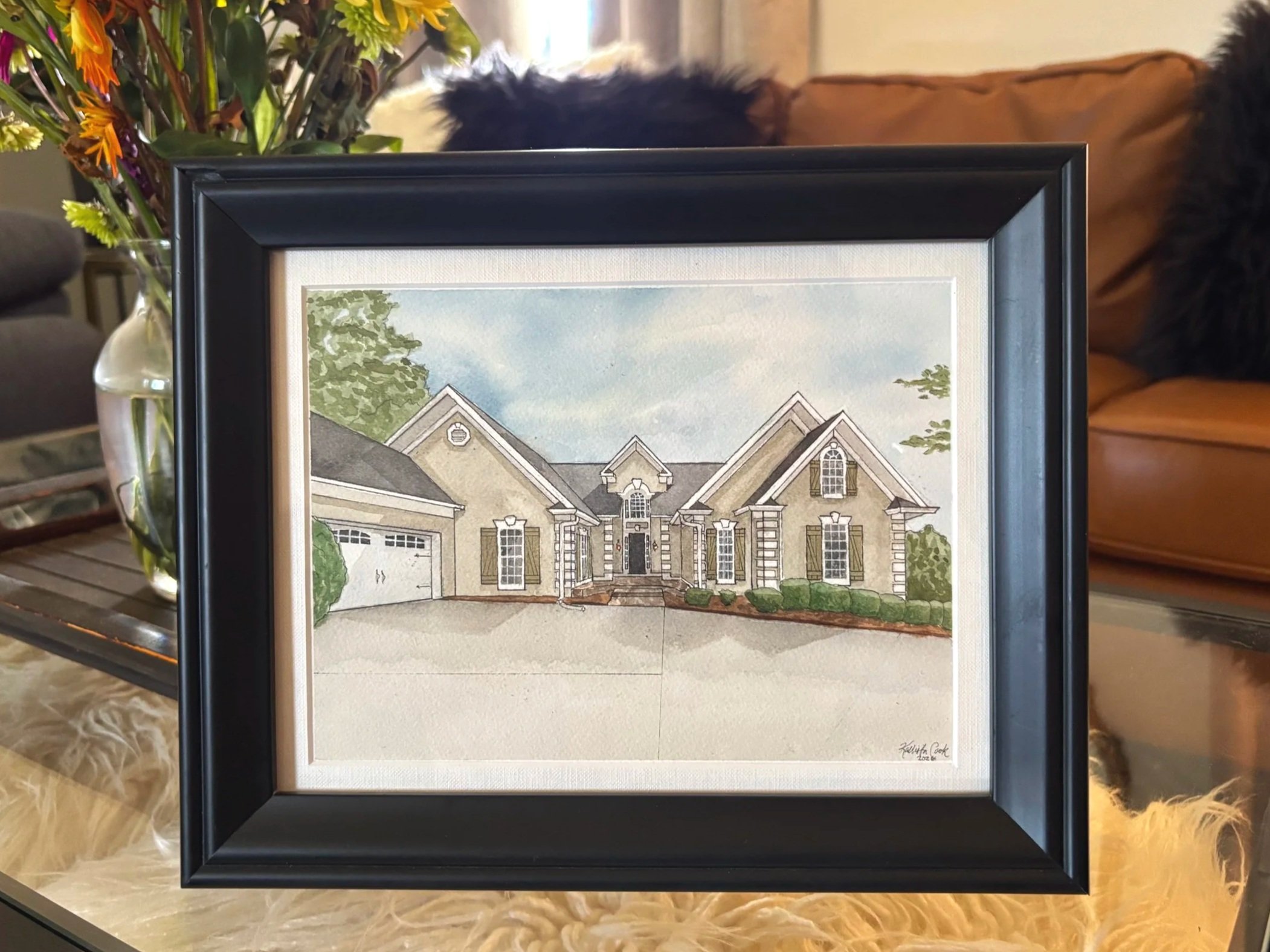 A framed watercolor painting of a large, modern house with a front yard, trees, and a cloudy sky in the background, placed on a glass table with a vase of flowers nearby.