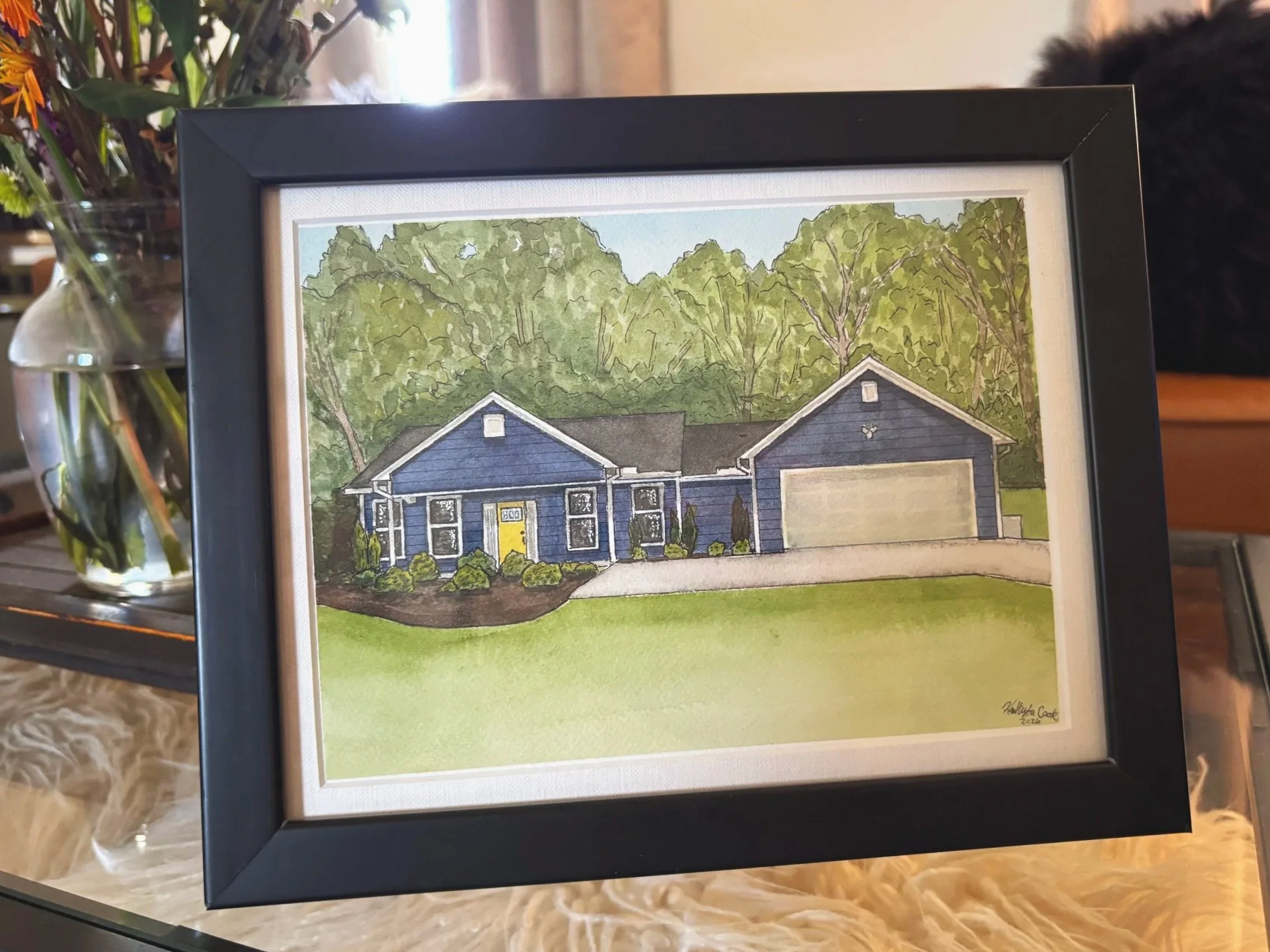 A framed watercolor painting of a blue house with a yellow door, surrounded by green trees and bushes, with a garage on the right and a manicured lawn in the foreground.