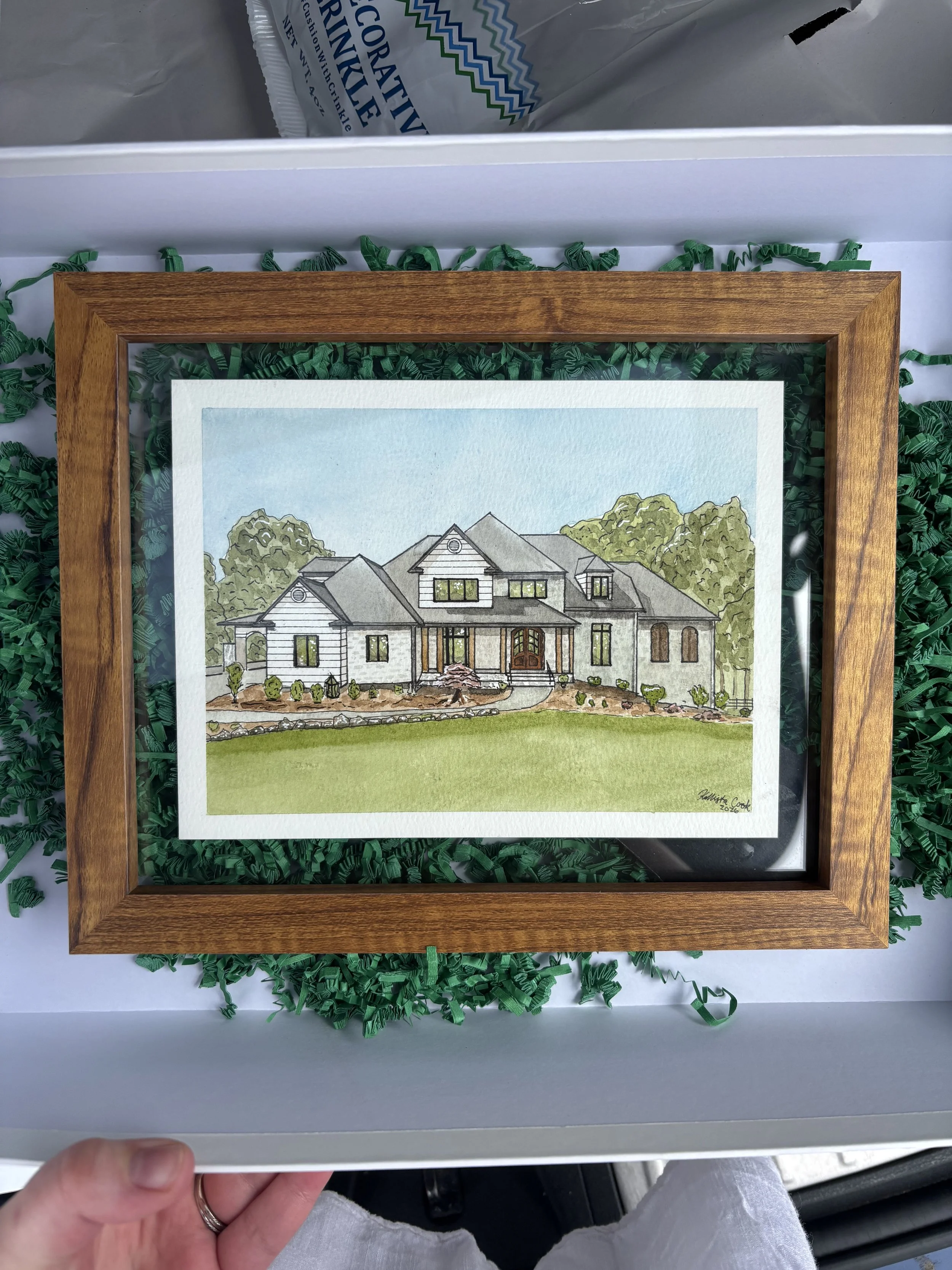 A framed watercolor painting of a large modern house with a landscaped front yard, set inside a white box with green crinkle paper.