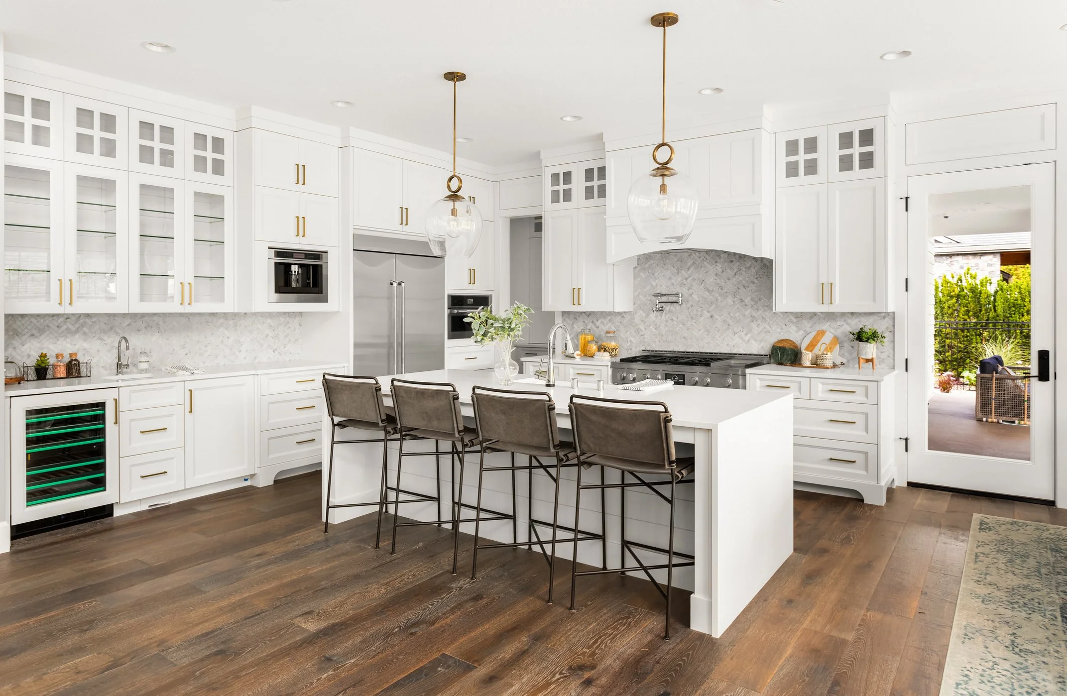 Modern white kitchen with a large island, gold hardware, glass-front cabinets, stainless steel appliances, pendant lights, and a sliding door leading to an outdoor patio.