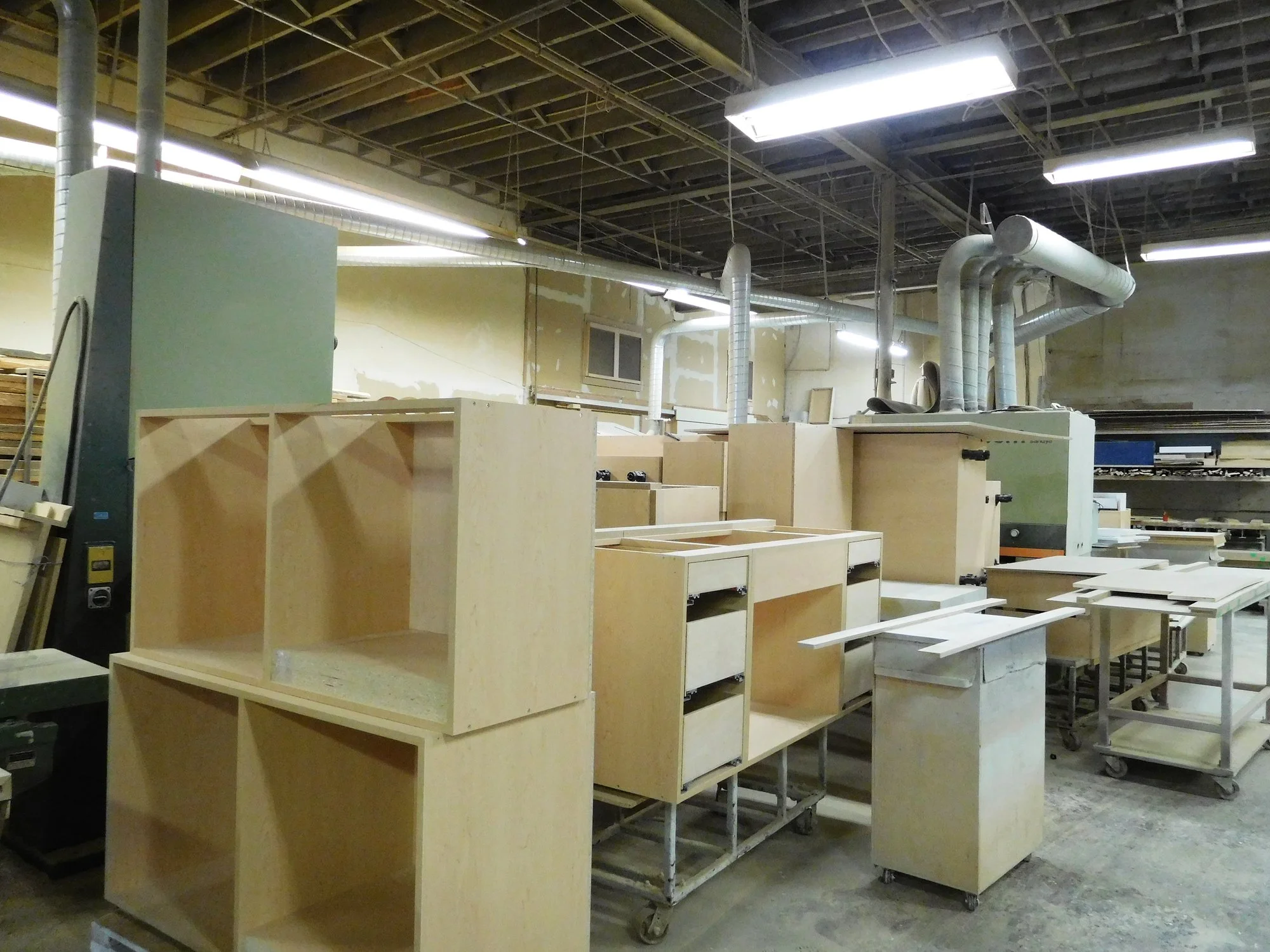 Wooden furniture pieces and cabinetry in an unfinished workshop or factory setting with exposed ceiling and ventilation pipes.
