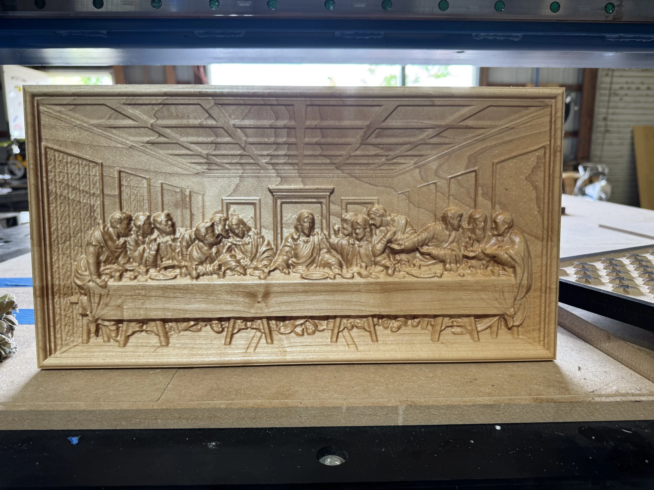 A detailed wooden carving of 'The Last Supper' scene, displayed on a wooden surface in a workshop setting with shelves and tools visible in the background.