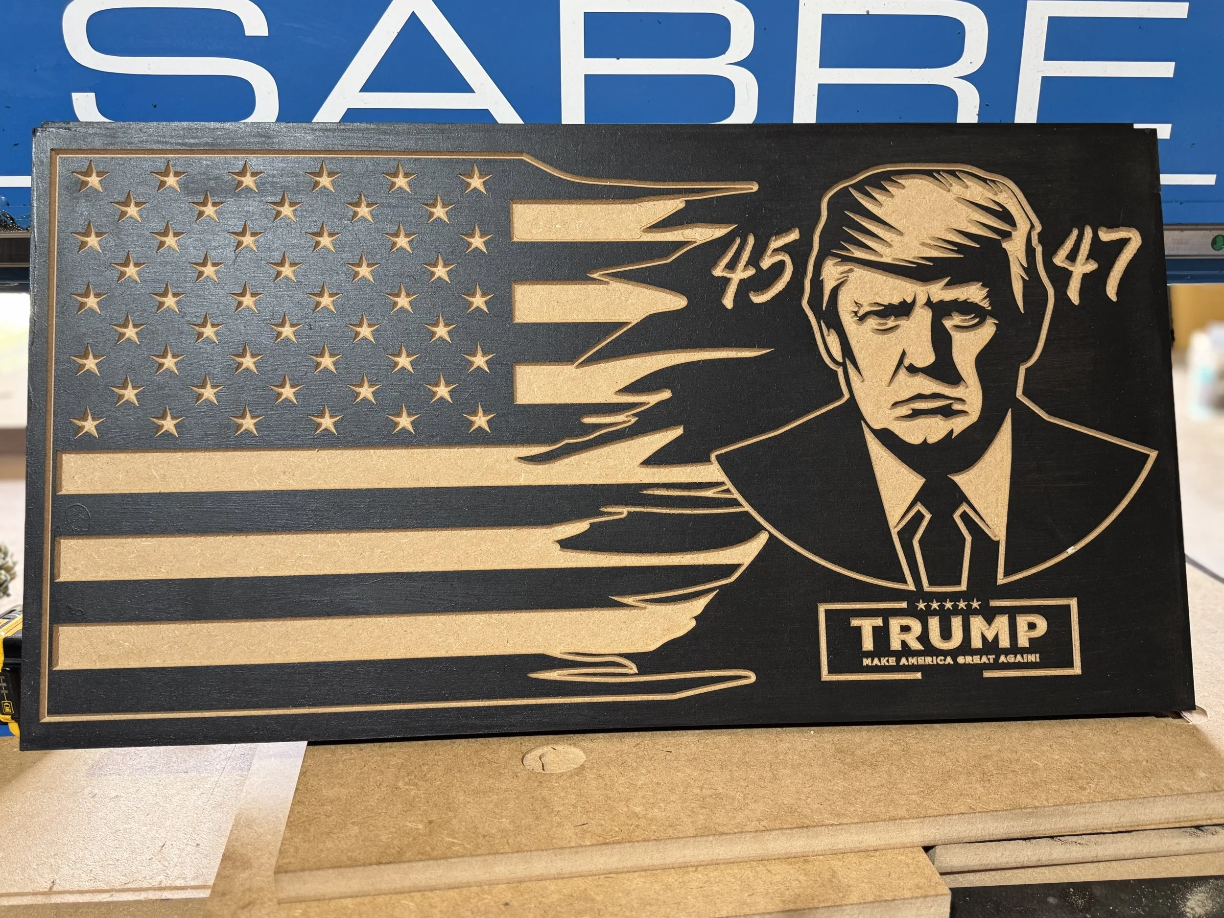 A metallic sign featuring the American flag with a stylized portrait of Donald Trump. The sign includes the text 'TRUMP' and 'MAKE AMERICA GREAT AGAIN!' at the bottom.