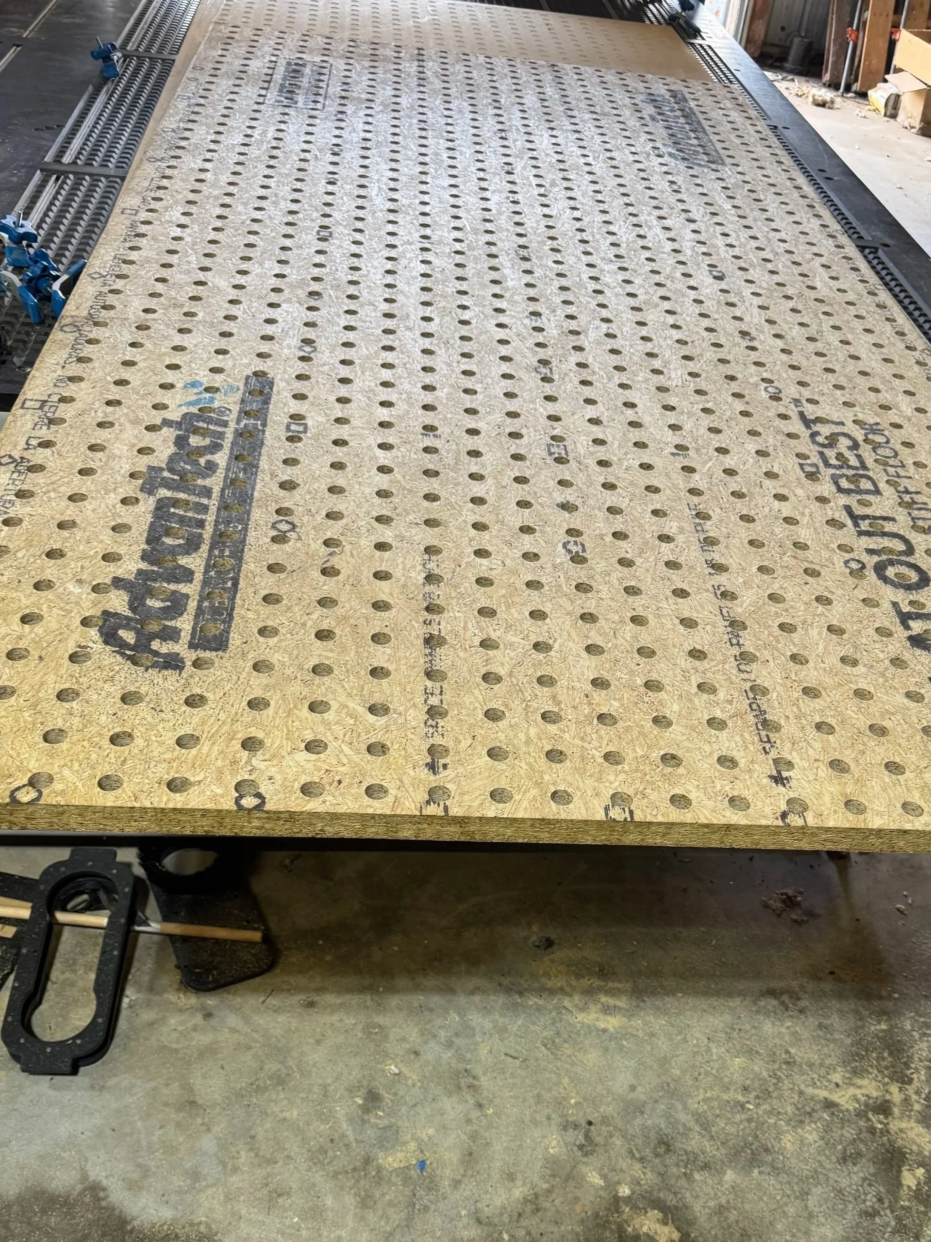 A workbench with a large sheet of perforated plywood on top, metal clamps on the side, and tools in a workshop environment.