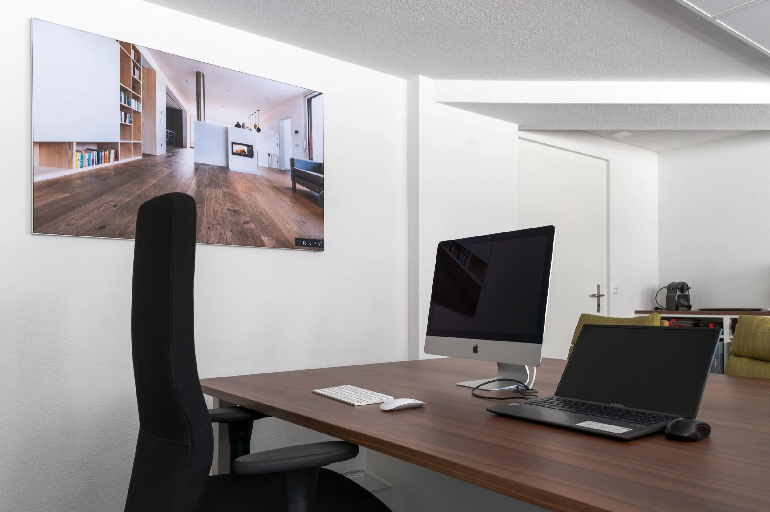 Ein modernes Büro mit einem Holzschreibtisch, einem iMac, einem Laptop, einer weißen Tastatur und einer Maus. An der Wand ist ein Fernseher oder Monitor, der ein Wohnzimmer mit Kamin und Holzboden zeigt.