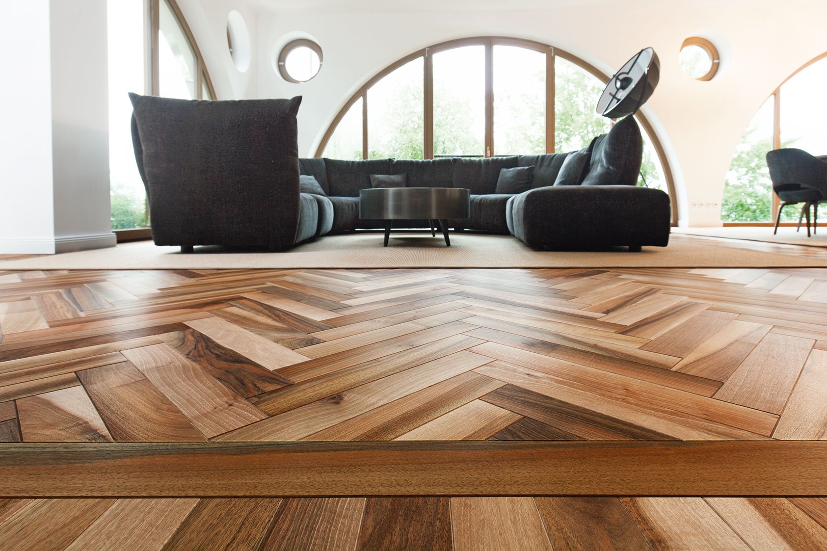 Modernes Wohnzimmer mit Hölzermosaikparkett, schwarzes Ecksofa, runder Couchtisch, große Fenster, runde Wandöffnungen, Stehleuchte, Wandlampe, Flügel-Sofa, moderne Einrichtung.