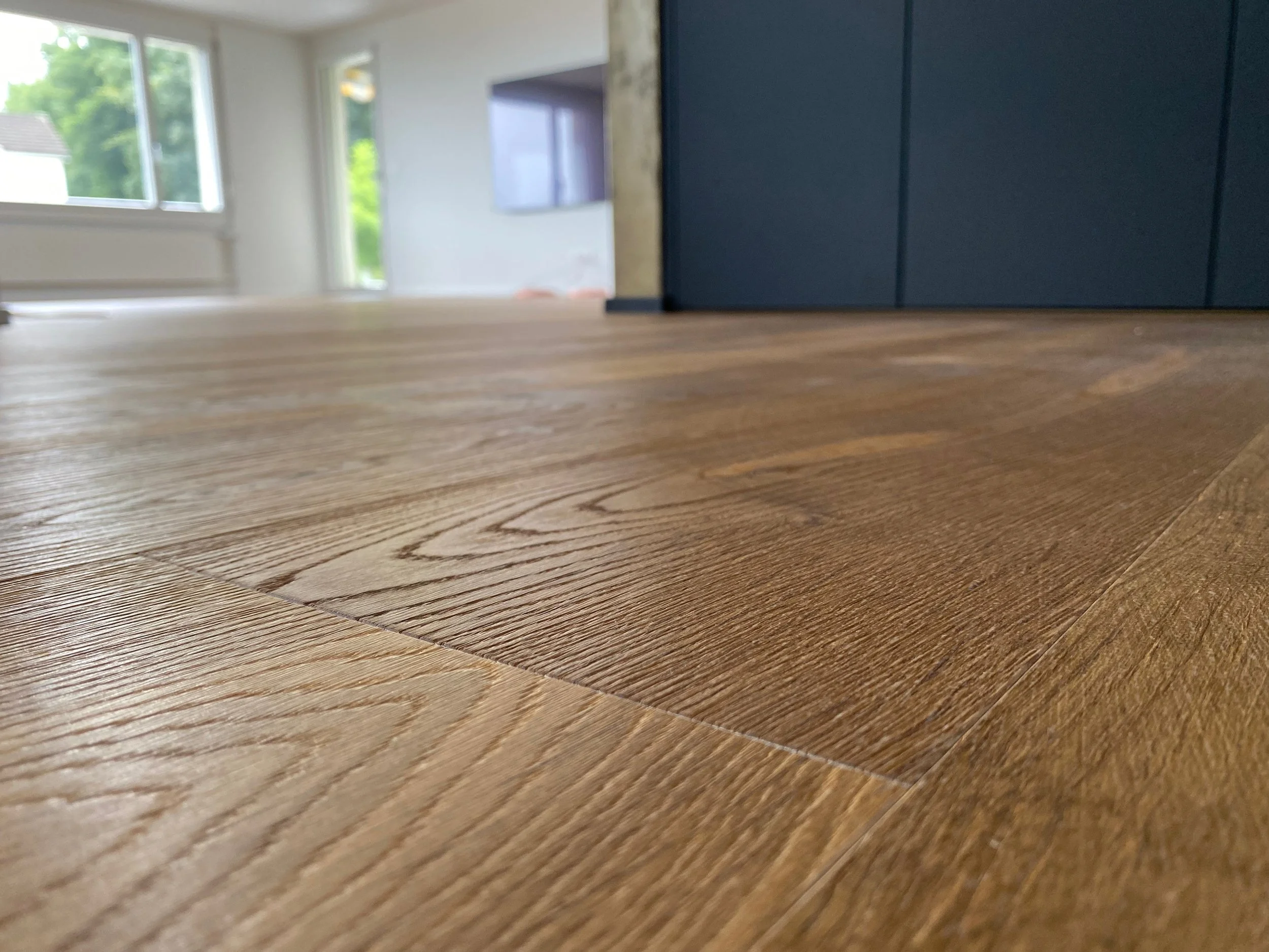 Blick auf Holzboden im Wohnzimmer mit weißen Wänden, Fenster und Fernseher im Hintergrund.