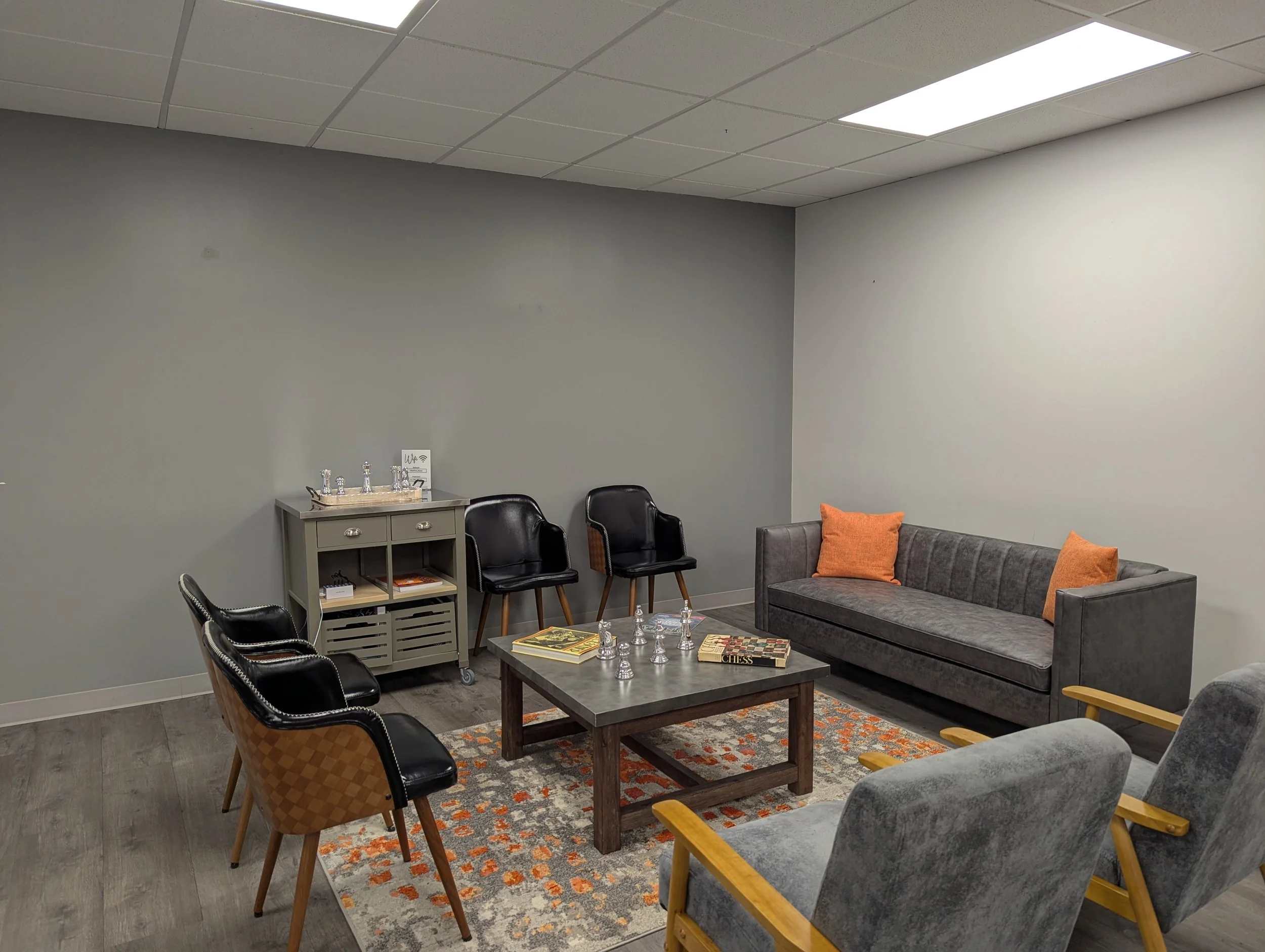 A waiting room or lounge area with gray walls, a gray ceiling with fluorescent lights, and gray flooring. There are multiple black chairs around a wooden coffee table with chessboards on it. A gray sofa with orange pillows is on the right side. A small gray table with a tray of water bottles and glasses, and some books, is against the wall. An orange rug and a few books are on the floor.