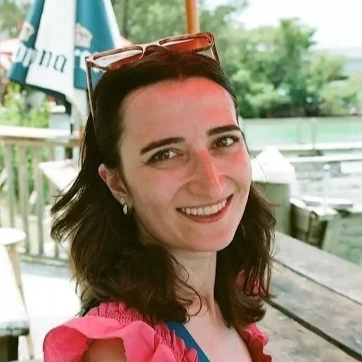 A woman with dark hair, wearing sunglasses on her head, smiling, in an outdoor setting with trees and a wooden railing.