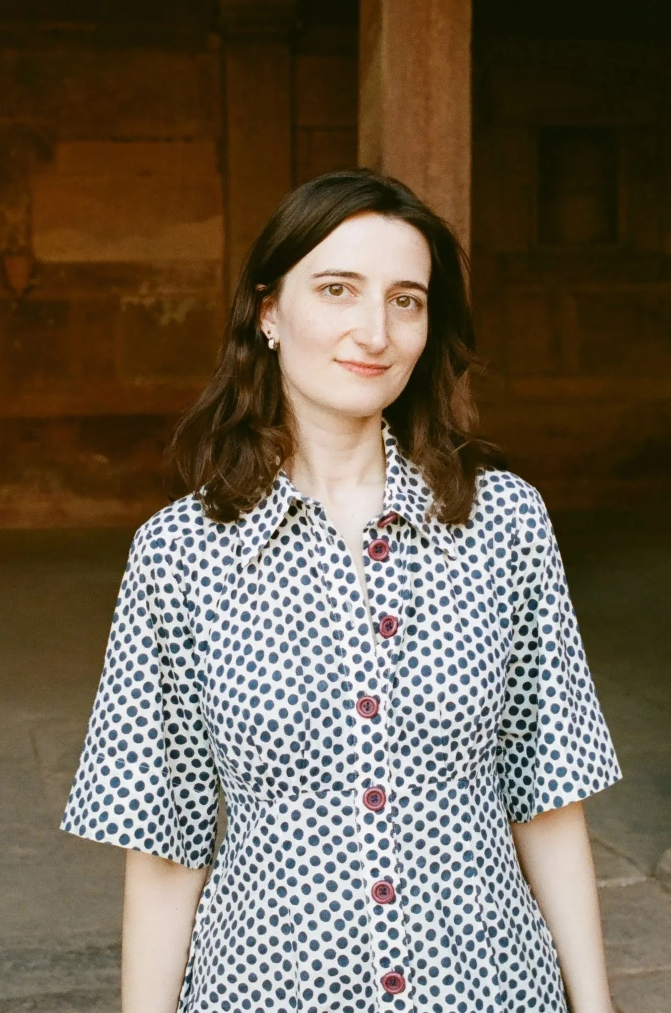 A woman with dark brown hair, light skin, wearing a white dress with navy polka dots and red buttons standing inside a wooden structure.