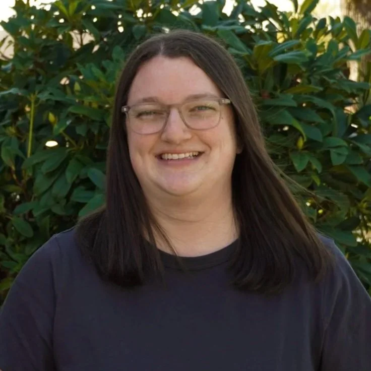A smiling woman with long dark hair and glasses, standing outdoors near a large tree with green leaves.