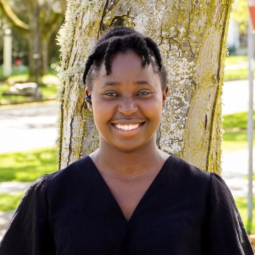 Young woman with styled hair, standing outdoors with trees and buildings in the background, smiling.