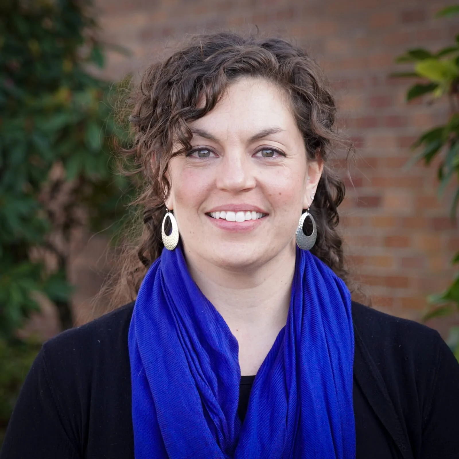 A woman with curly brown hair, wearing large silver earrings, a black top, and a bright blue scarf, smiling outdoors in front of green foliage and a brick wall.