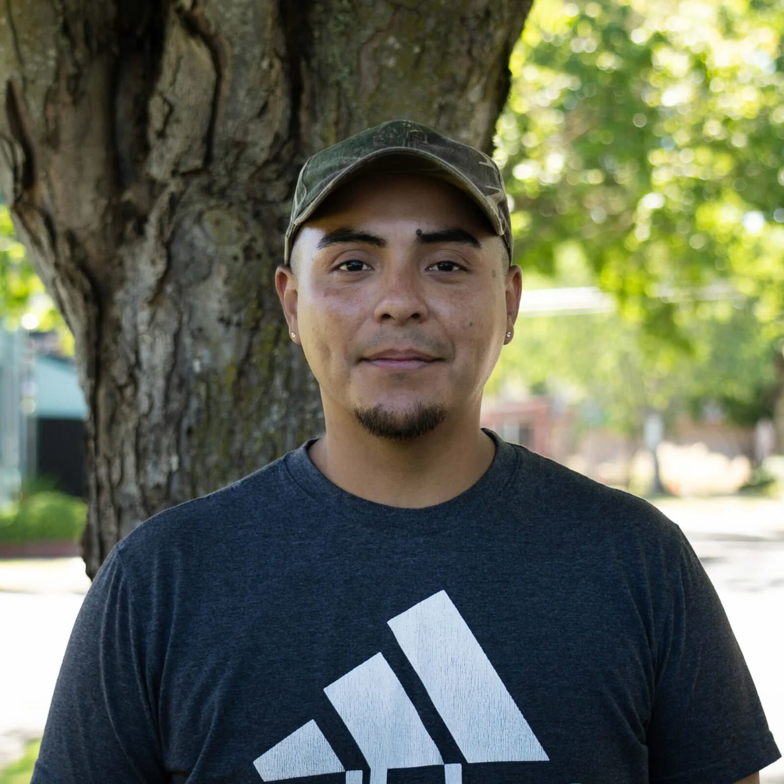 A man standing outdoors in front of a large tree with green leaves, wearing a dark gray t-shirt with a white geometric design and a camouflage baseball cap.