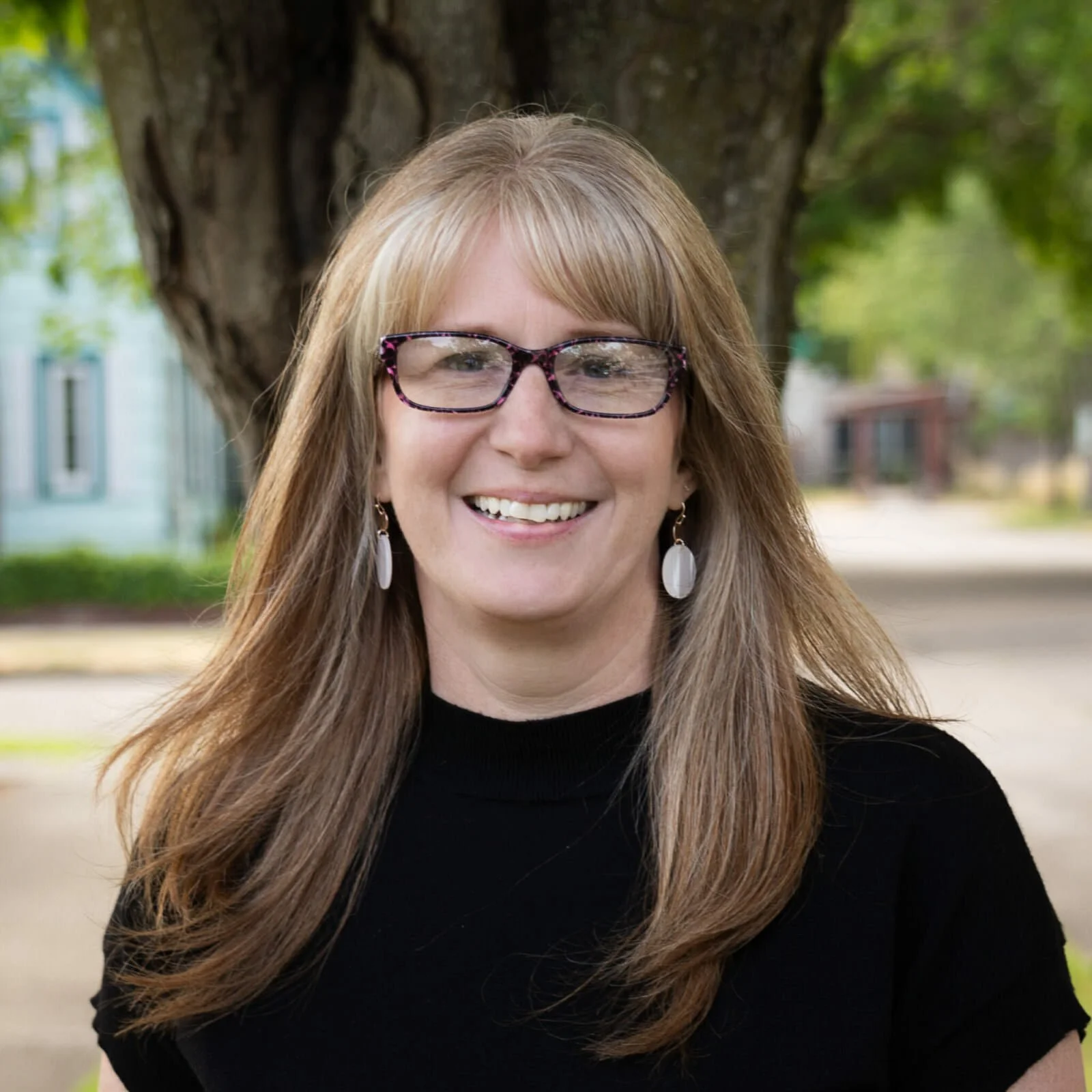 A woman with long light brown hair, wearing glasses with a floral pattern, earrings, and a black top, smiling outdoors near a large tree.
