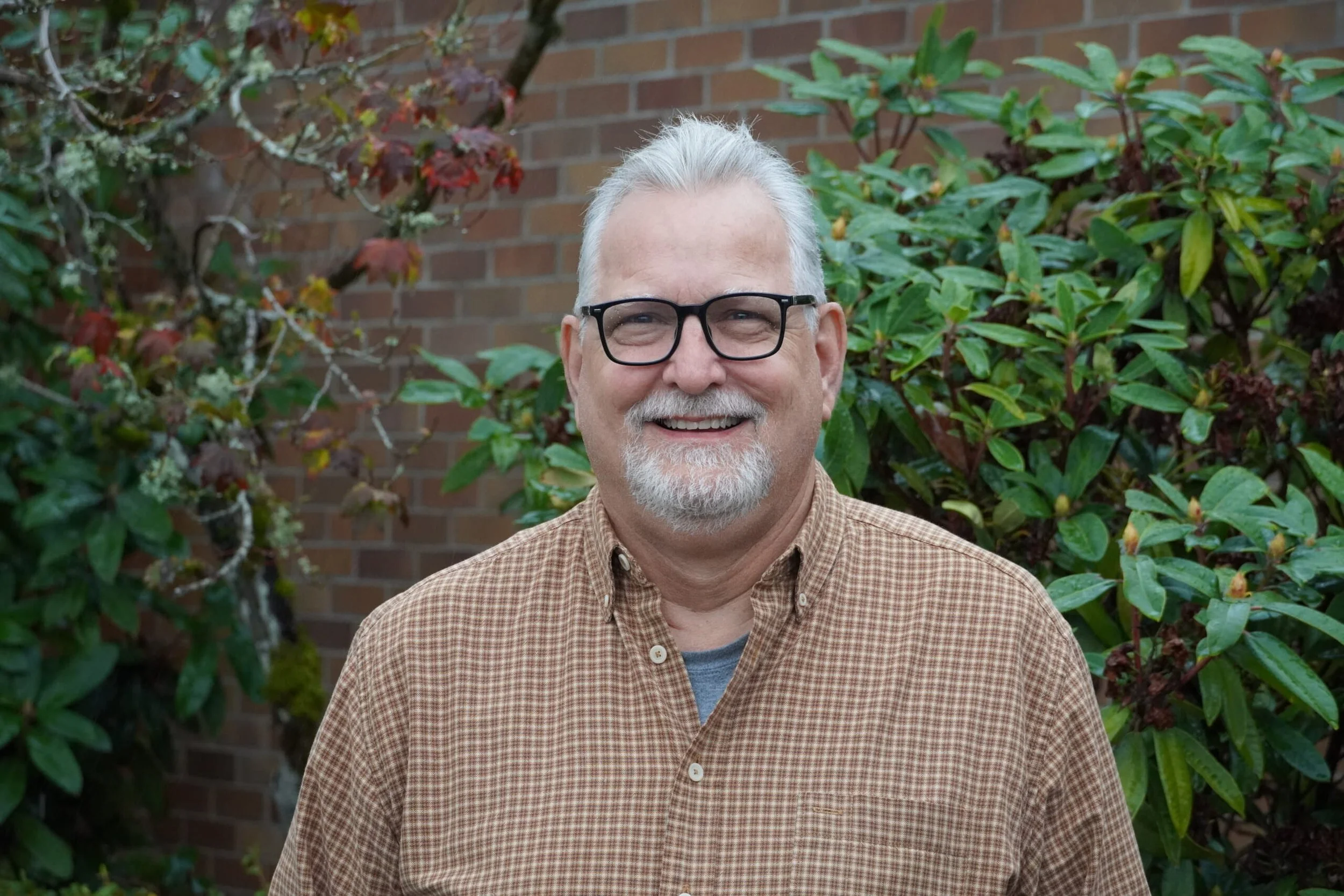 A smiling middle-aged man with gray hair, beard, and glasses stands outdoors in front of green shrubbery and a brick wall.