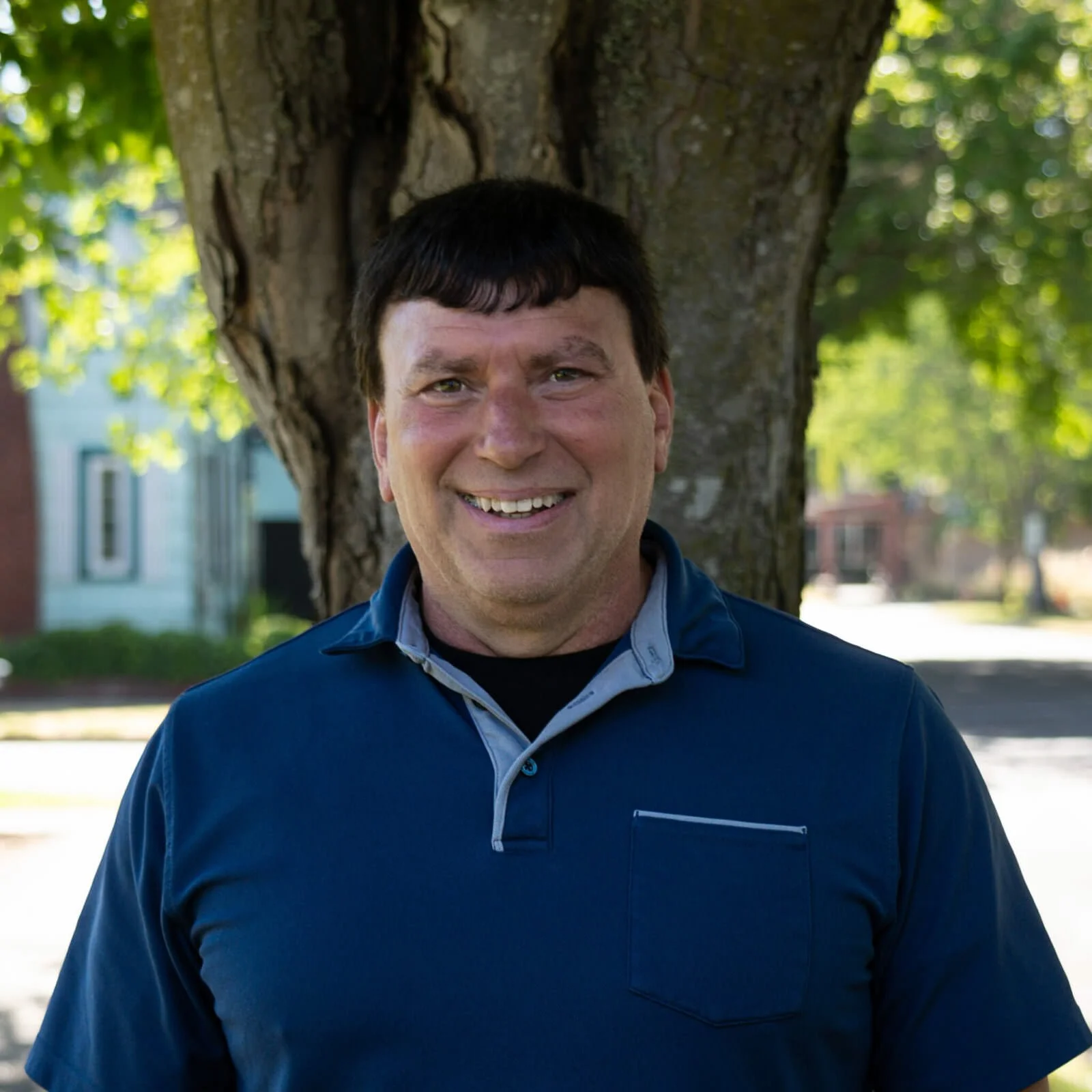A man smiling outdoors standing in front of a tree with green leaves, wearing a blue collared shirt.
