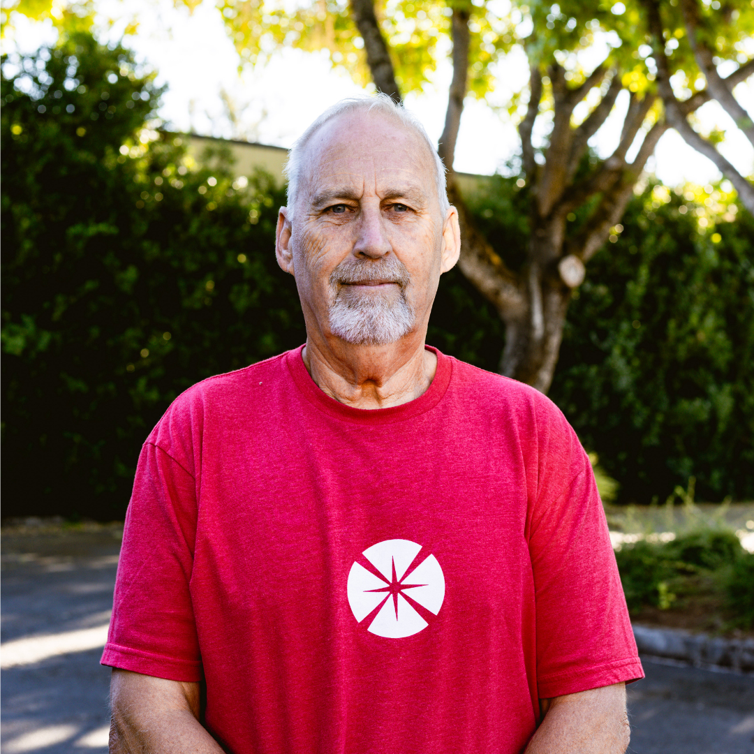 An elderly man with white hair and a beard standing outdoors on a sunny day, wearing a red t-shirt with a white circular symbol on it, surrounded by trees and greenery.