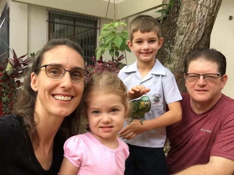A family of four posing outdoors with a bird, in front of a tree and house. The woman and girl are in the front, the boy and man are behind them. The boy holds the bird, and everyone is smiling.