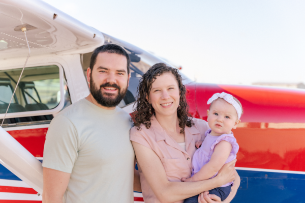 Family of three standing in front of small airplane, smiling, outdoors, clear day