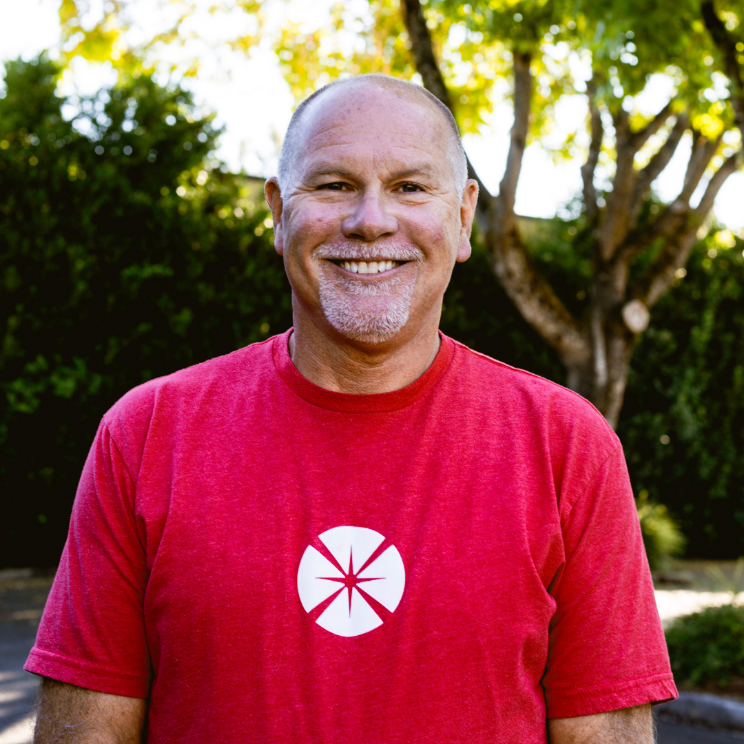 Smiling middle-aged man with a goatee and bald head wearing a red t-shirt with a white symbol, standing outdoors with trees and sunlight in the background.
