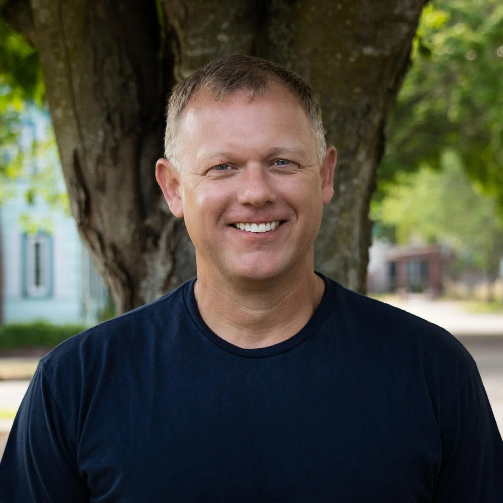 A man wearing glasses and a blue button-up shirt, standing outdoors in front of a large tree and greenery, smiling at the camera.