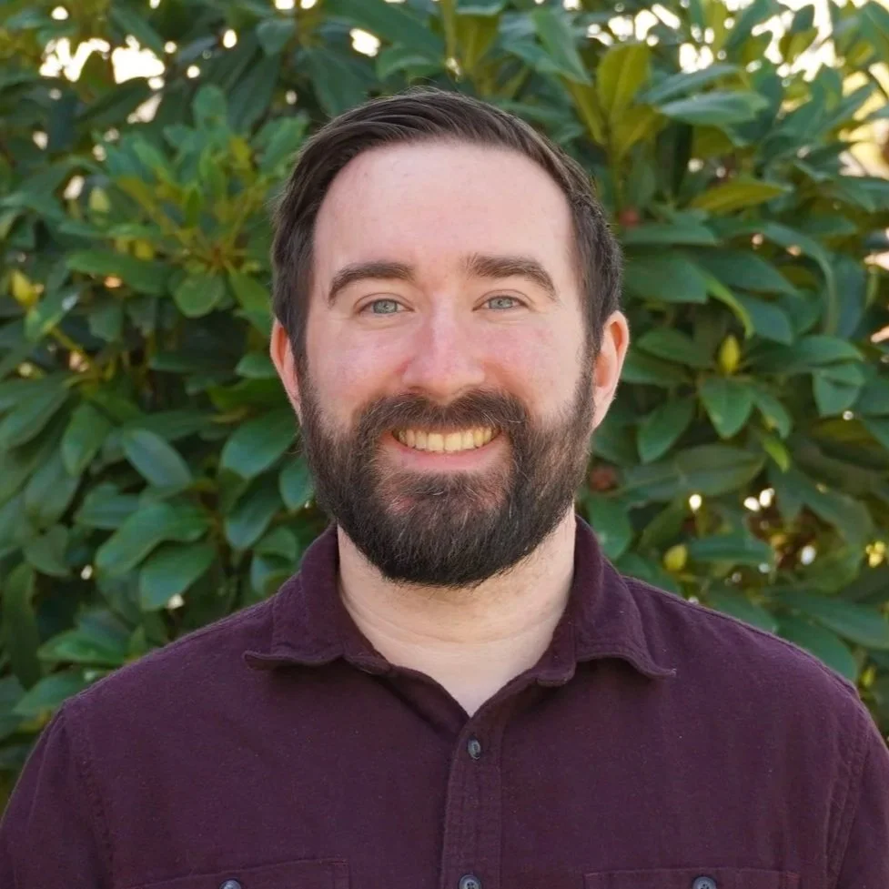 A man with dark hair, a full beard, and light skin smiling outdoors in front of a large tree with green leaves, wearing a burgundy shirt.