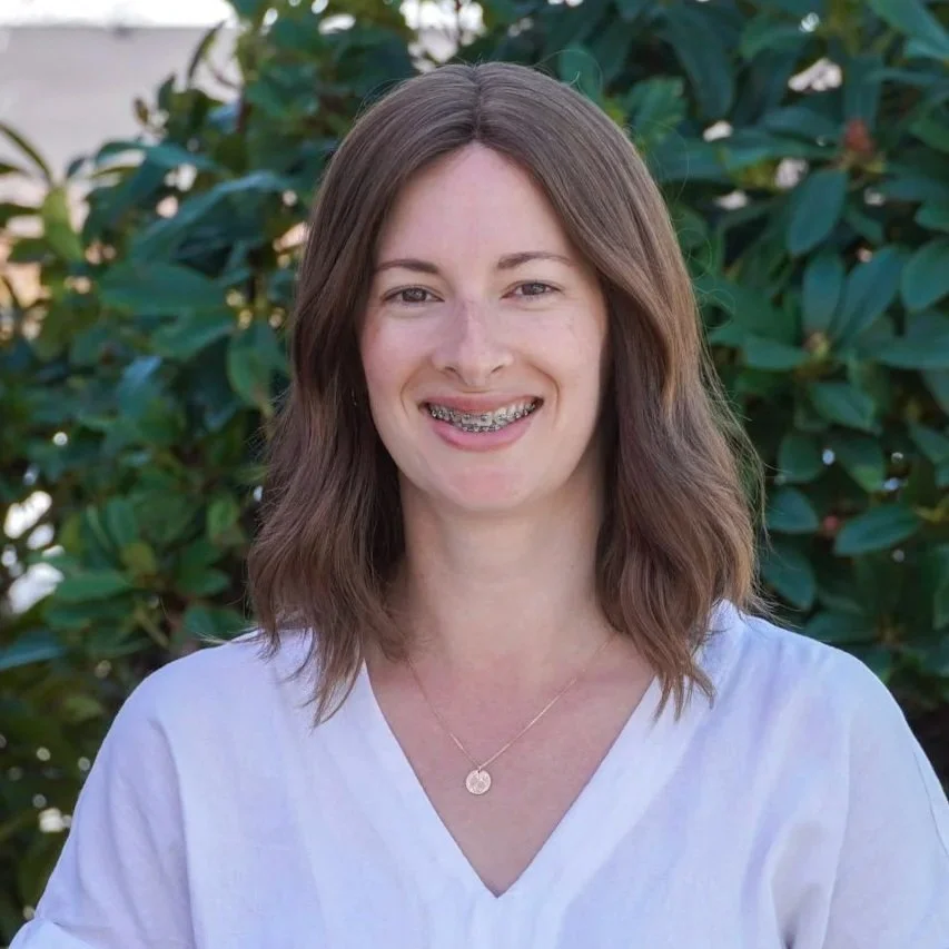 A woman smiling outdoors with trees and a building in the background.