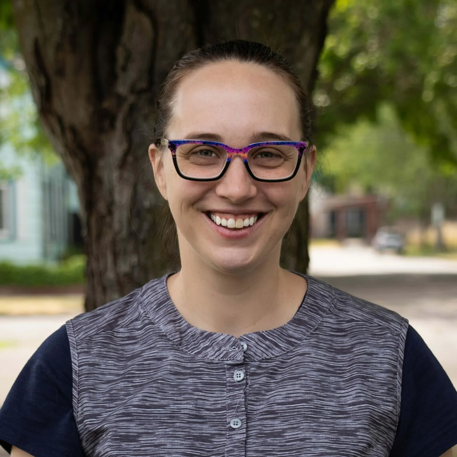A woman smiling outdoors with a tree behind her, wearing glasses and a striped shirt.