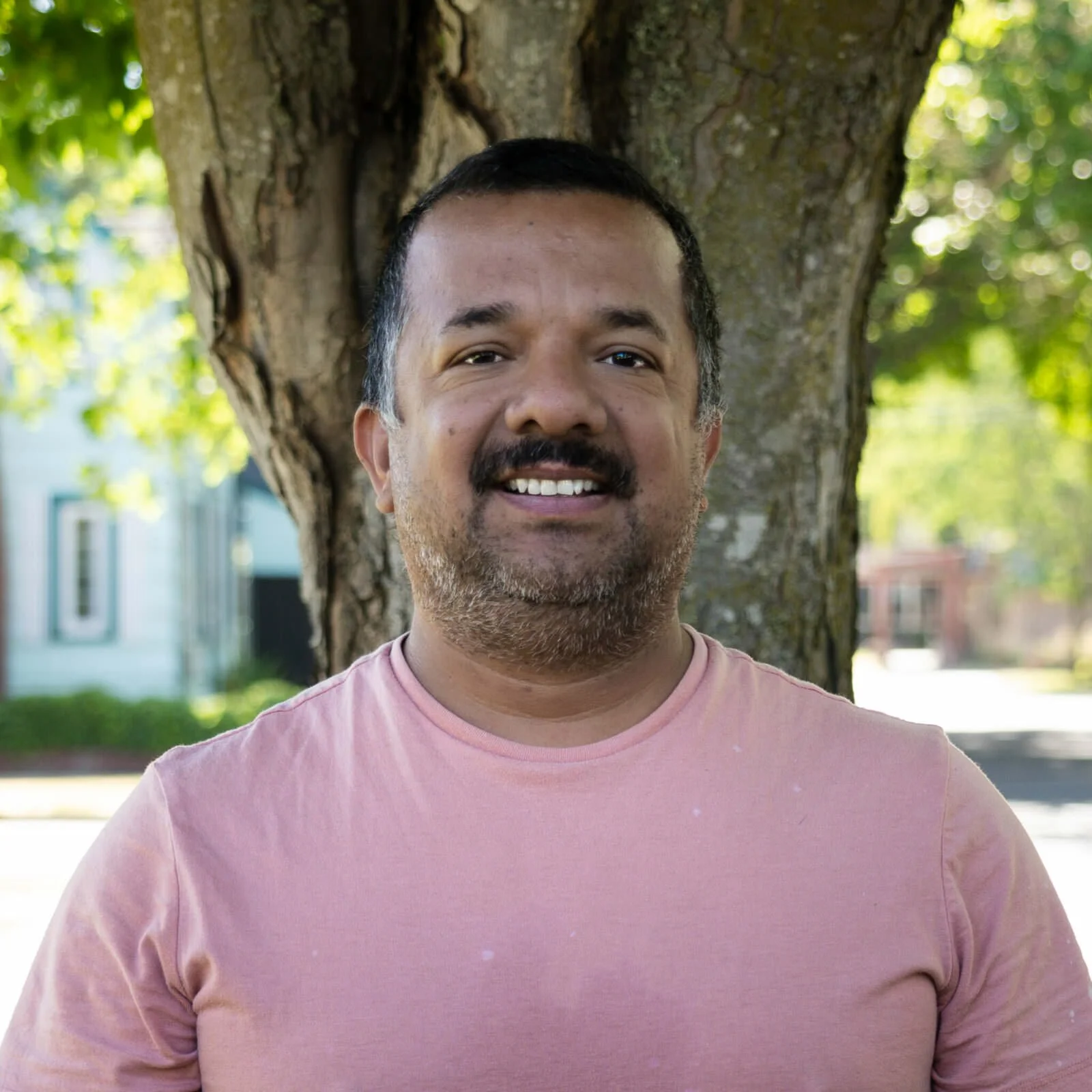 A smiling man with short dark hair and a beard standing outdoors in front of a tree with green leaves, wearing a pink t-shirt.
