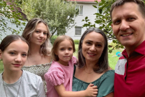 A family of five posing together outdoors in a garden with trees and a house in the background. The group includes two young women, a young girl, a woman, and a man.