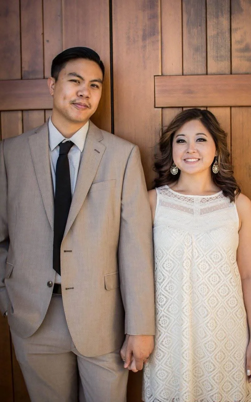 A man and a woman standing and holding hands, leaning against a wooden wall. The man is wearing a beige suit with a black tie, and the woman is wearing a white dress with lace detail and earrings.