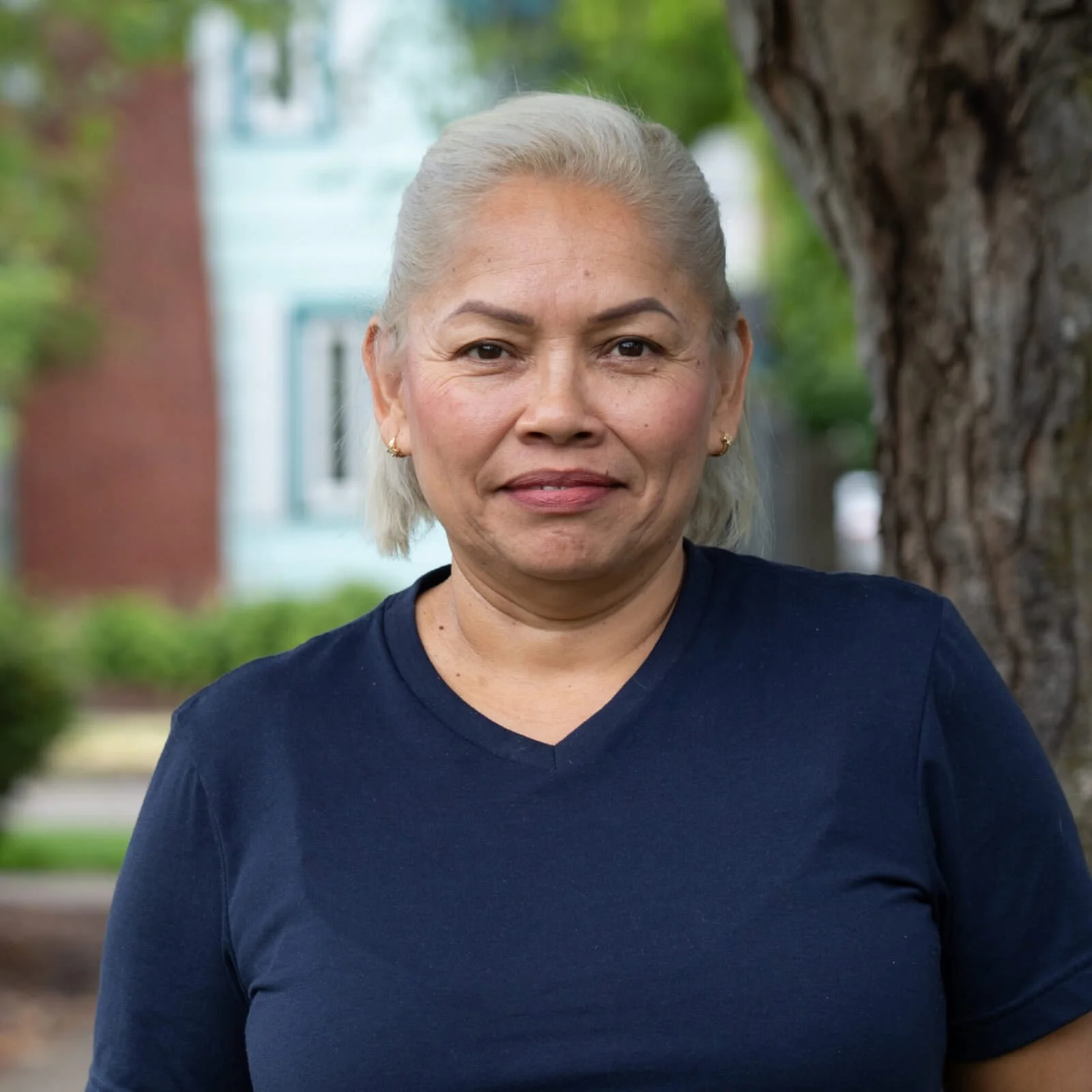 An older woman with blonde hair wearing a navy blue top standing outdoors near a large tree with a blurred background of greenery and buildings.