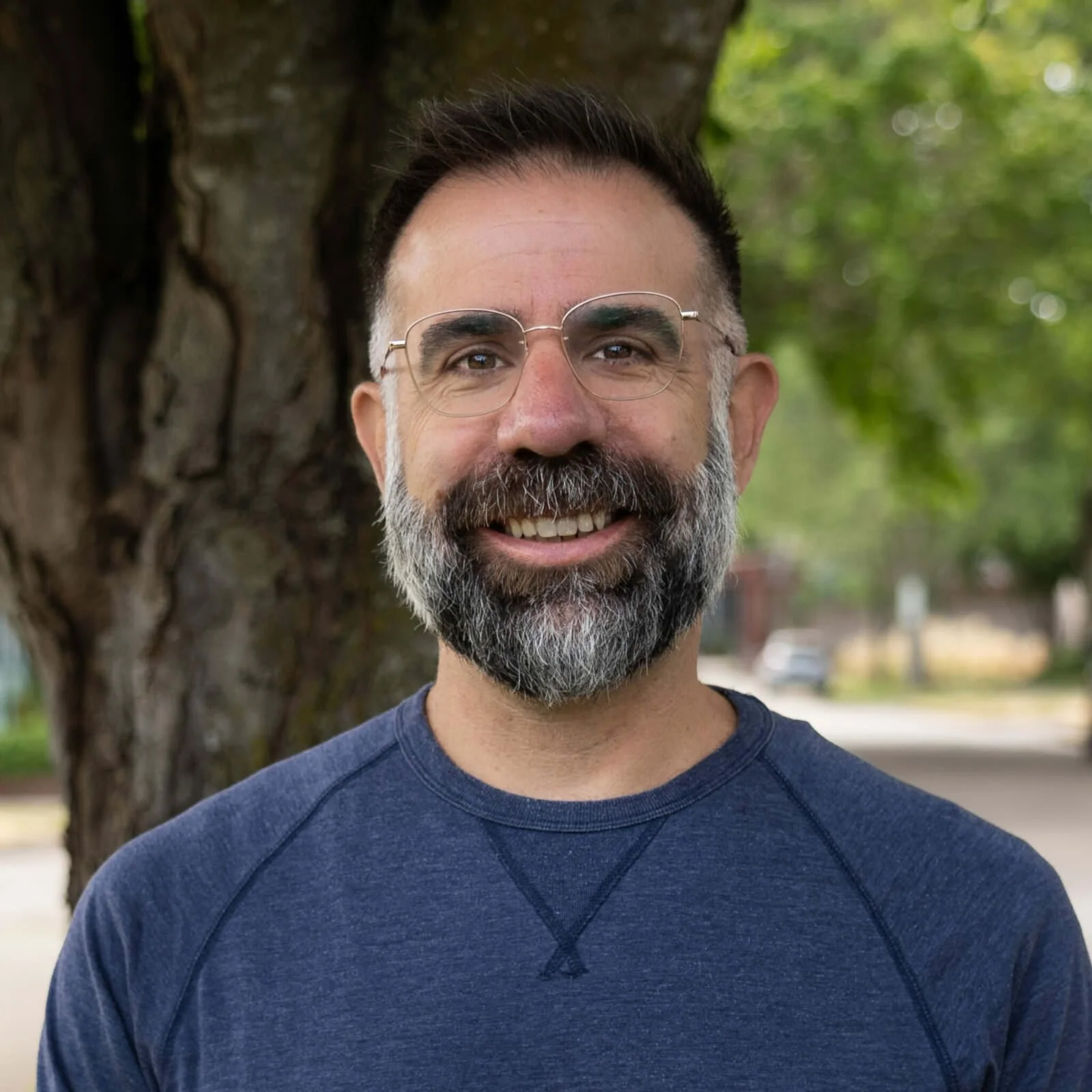 Smiling man with glasses and a beard outdoors near a tree.