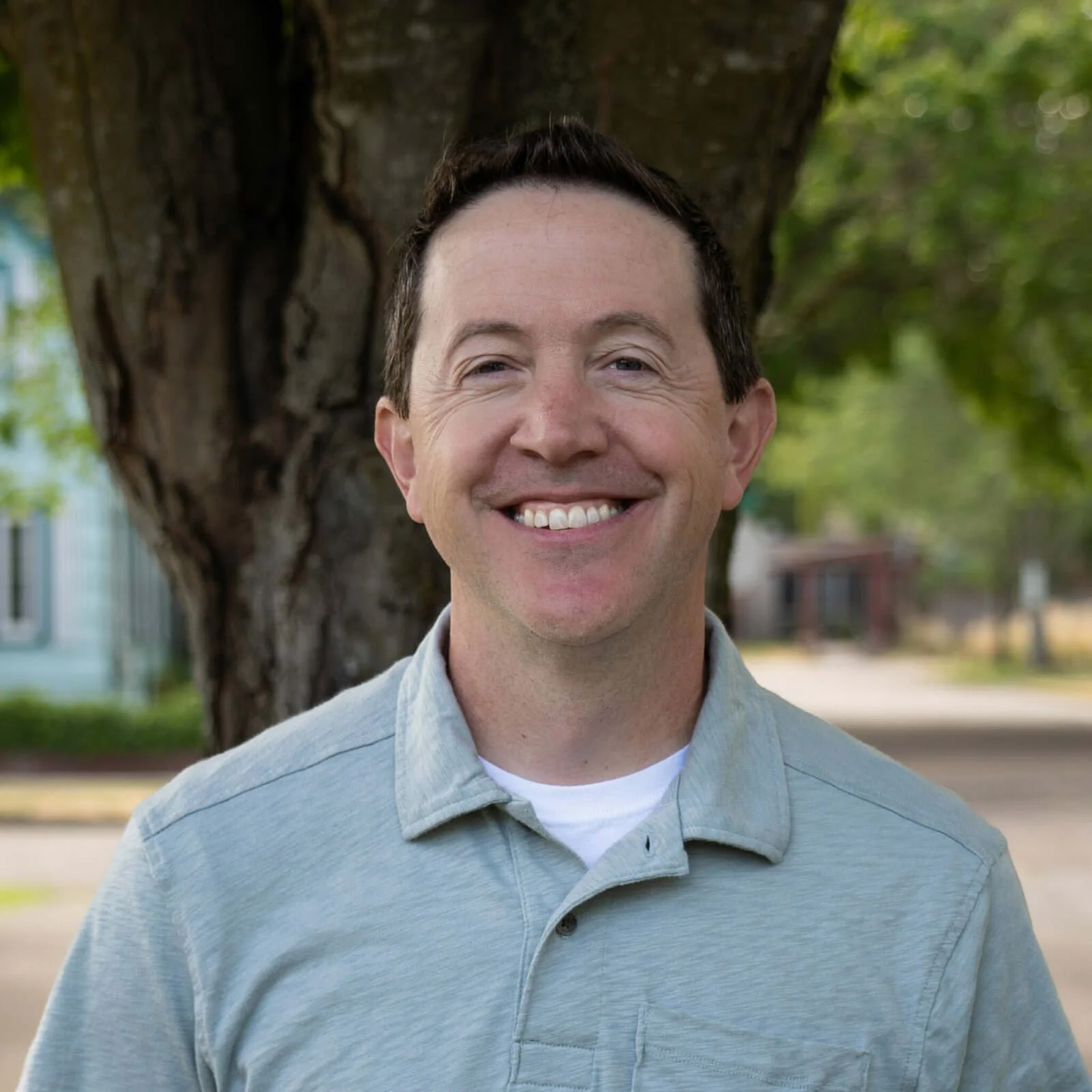 A man smiling outdoors standing in front of a tree with green leaves, wearing a blue collared shirt.