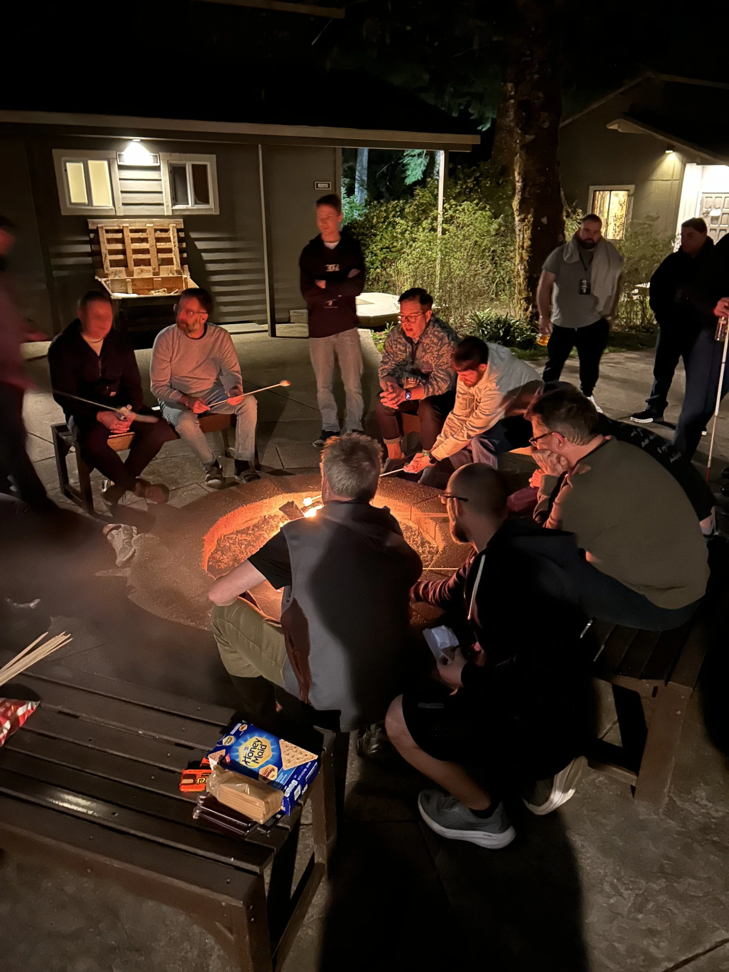 Group of people sitting around a fire pit outdoors at night, roasting marshmallows, with some standing nearby.