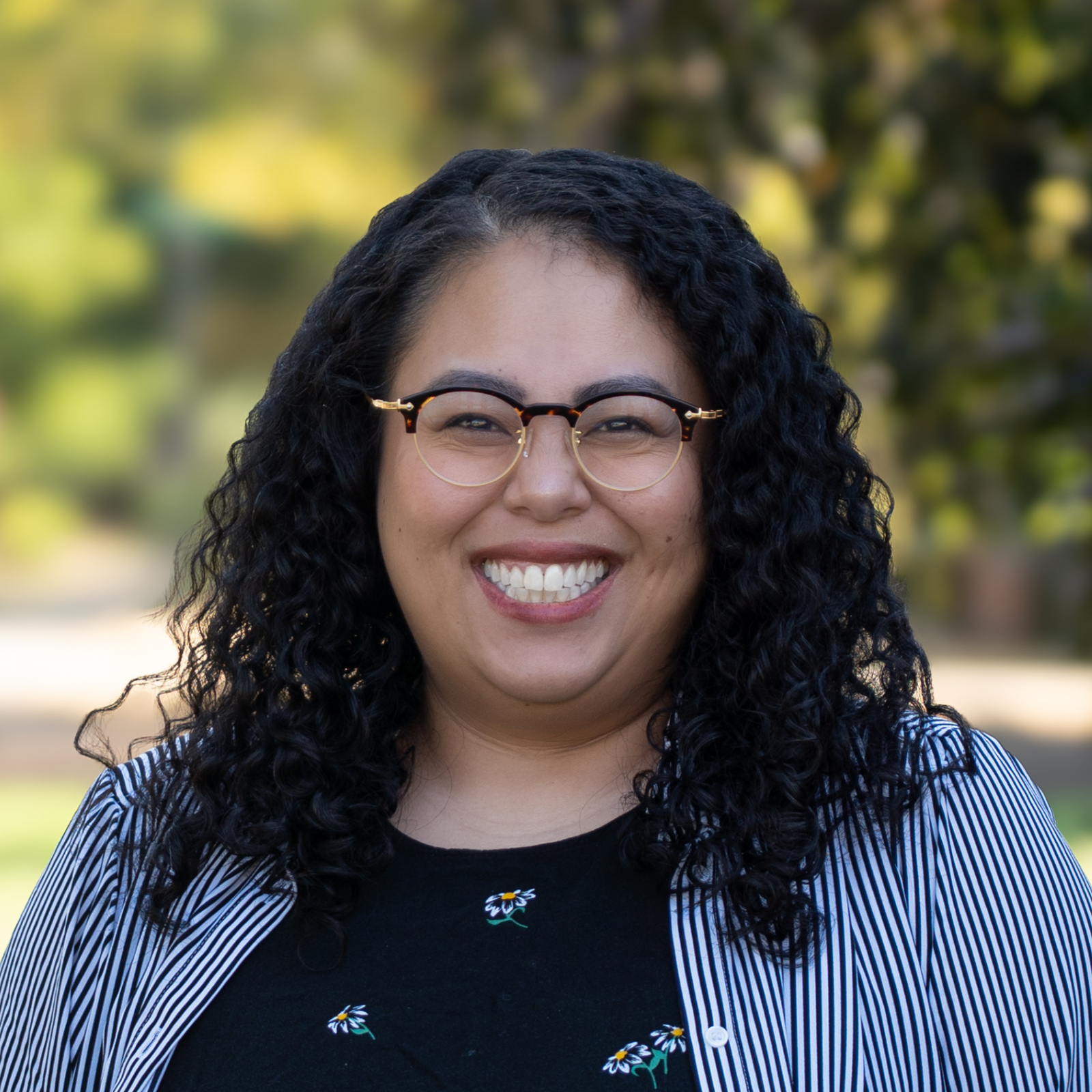 A smiling woman with curly black hair, wearing glasses and a striped blazer, standing outdoors with blurred trees and greenery in the background.