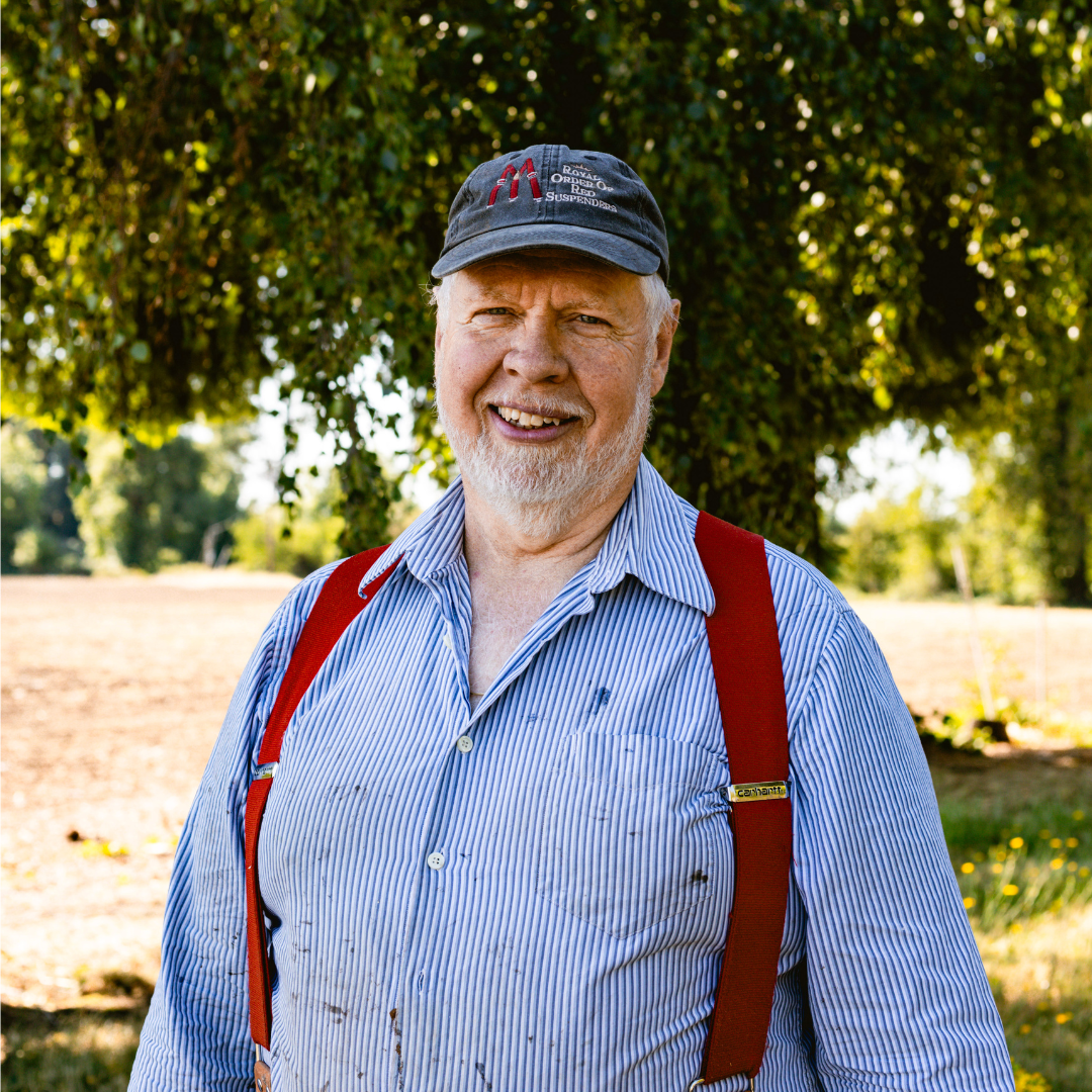 An elderly man with a gray beard wearing a black baseball cap, a blue striped shirt, and red suspenders, standing outdoors near a tree with green leaves in the background.