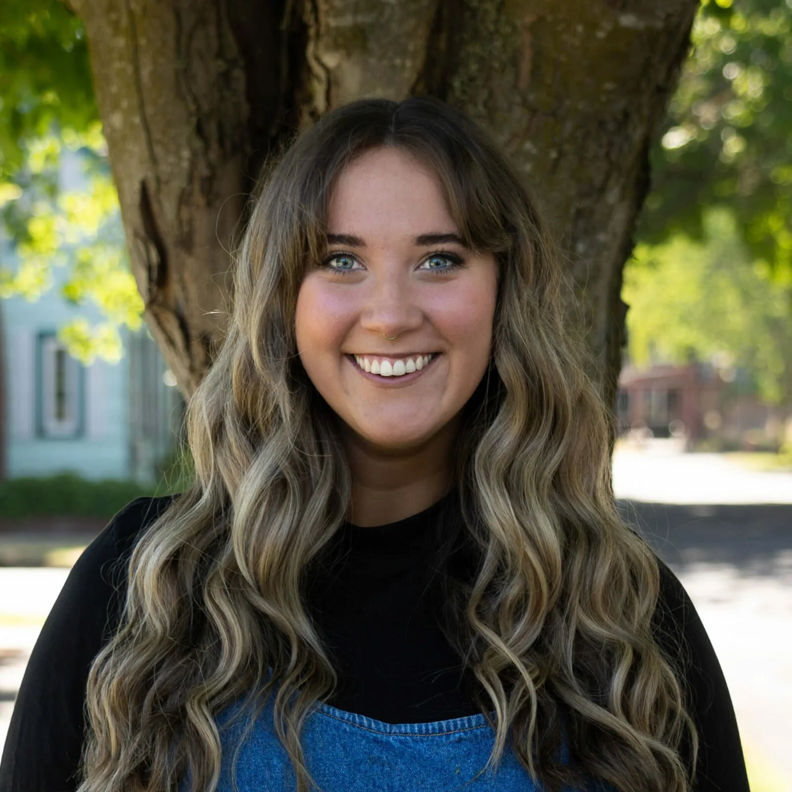A young woman with long wavy hair, wearing a black shirt and denim overalls, smiling outdoors in front of a large tree with green leaves.