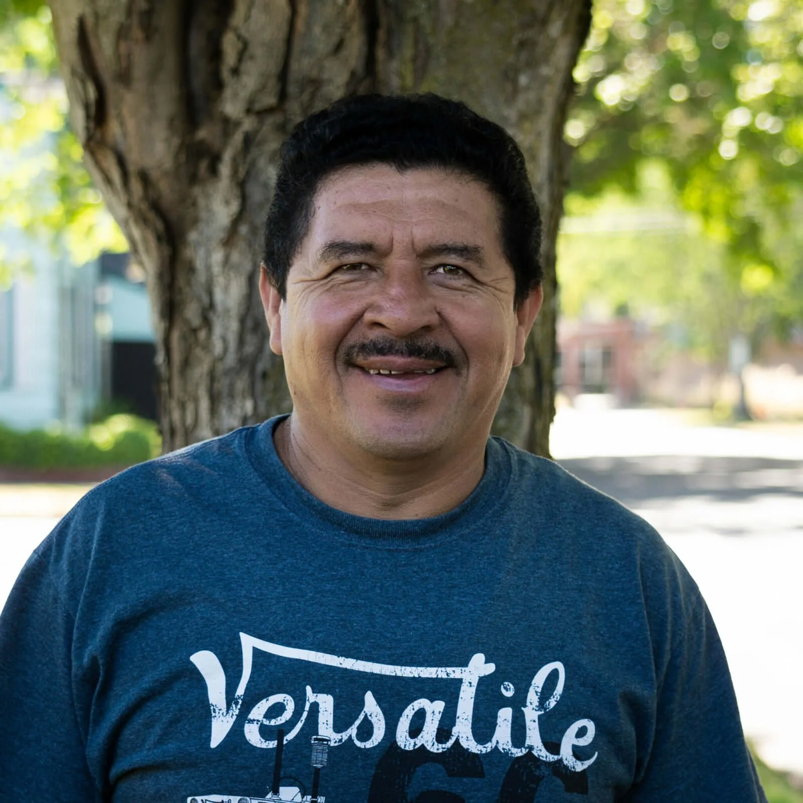 A middle-aged man with dark hair, a mustache, and a friendly smile, standing outdoors in front of a tree with green leaves. He is wearing a blue t-shirt that says 'Versatile'.