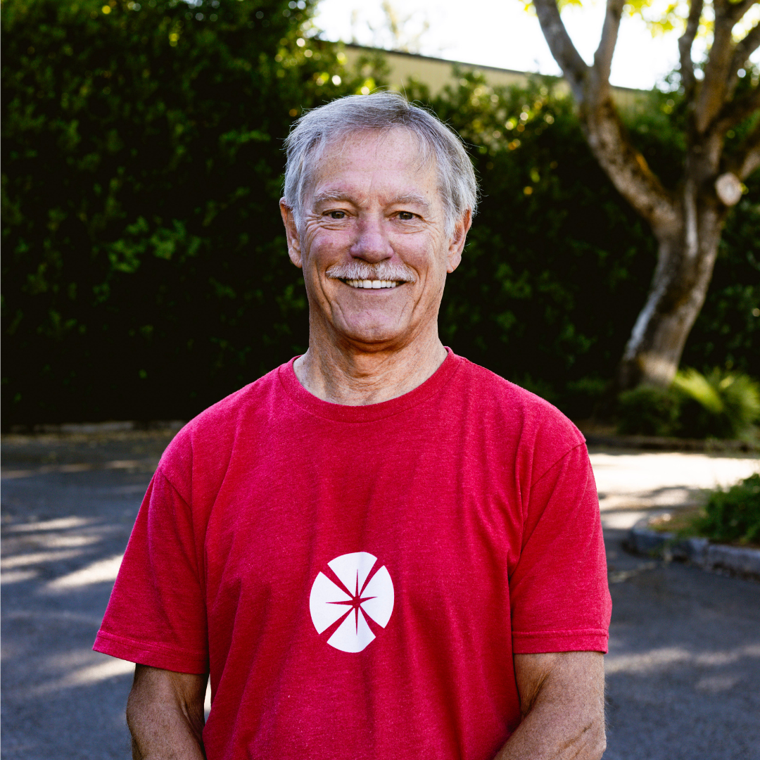 Smiling elderly man standing outdoors in a red T-shirt with a white circular logo, trees and shadows in the background.